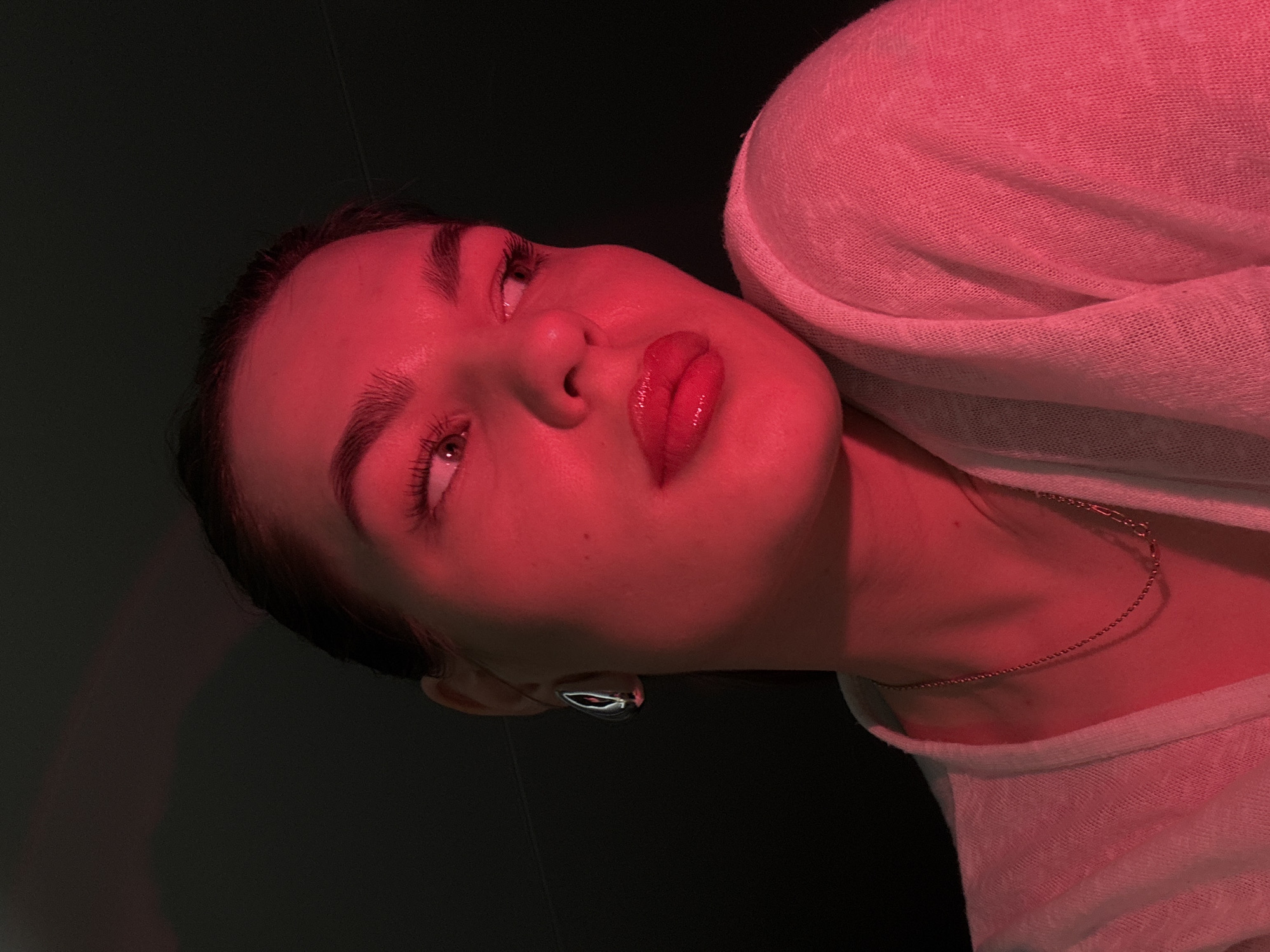 A low-angle close-up photograph captures a woman with light skin bathed in red light. She is wearing a light-colored, possibly white, knit top and a delicate gold chain necklace. She also has a silver teardrop earring visible on her left ear. The background is dark, appearing to be a plain wall. There's no information in the image to confirm it was taken in the city of Chaikovsky, Russia.