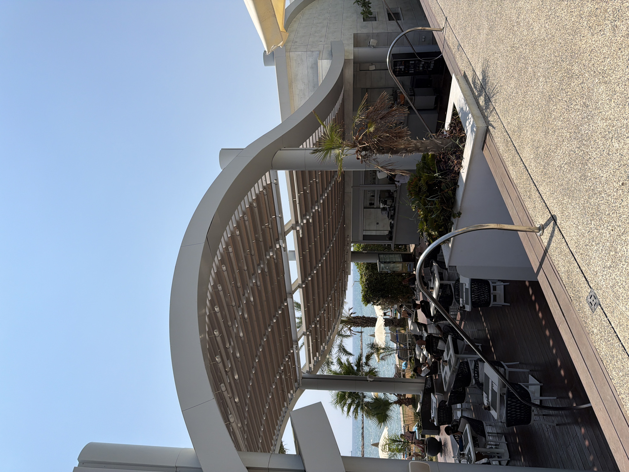 An outdoor dining area is set up with tables and chairs beneath a modern, wave-like canopy structure. Beyond the canopy, a view of the sea is visible, with lounge chairs and umbrellas on a beach. Palm trees are scattered throughout the scene, adding to the tropical ambiance. The architecture features clean lines and a predominantly white and grey color palette. The setting appears to be a restaurant or cafe located in Oroklini, Cyprus, given the coastal scenery and clear blue sky. There are several patrons seated at tables, some engaged in conversation and others appearing to relax. The overall atmosphere suggests a leisurely daytime setting at a seaside establishment.