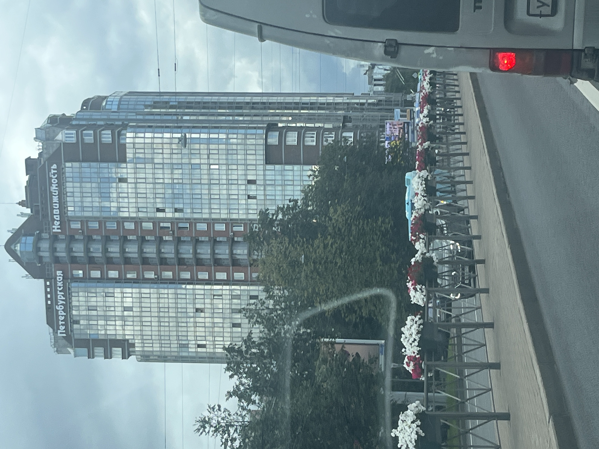 The image depicts a view from a vehicle, likely a van, looking towards a tall, modern apartment building in Saint Petersburg, Russia. The building features a facade of glass and reddish-brown brickwork. The text "Петербургская" (Peterburgskaya) and "Недвижимость" (Nedvizhimost) are visible on the upper portion of the building, indicating it is related to St. Petersburg real estate. In the foreground, a row of planters filled with white and red flowers is visible along a metal railing, bordering a sidewalk and a street. The sky is overcast, suggesting cloudy weather. The scene appears to be a street view in an urban environment.