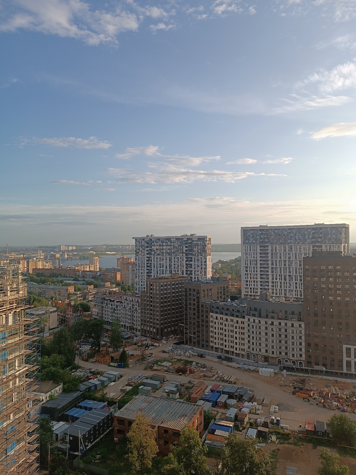 This is an aerial, eye-level, medium shot of a city under a bright, cloudy sky. In the foreground, construction is underway on a large residential building with scaffolding covering its exterior. Adjacent to this building, a construction site is visible with numerous modular units, building materials, and construction equipment. Further into the scene, several completed apartment buildings of varying heights and architectural styles dominate the urban landscape. In the background, a body of water, possibly a river or lake, separates the city from a distant, low-lying landscape with more buildings. The overall impression is one of ongoing development and urban expansion. The location is the city of Idrisovo, Russia.