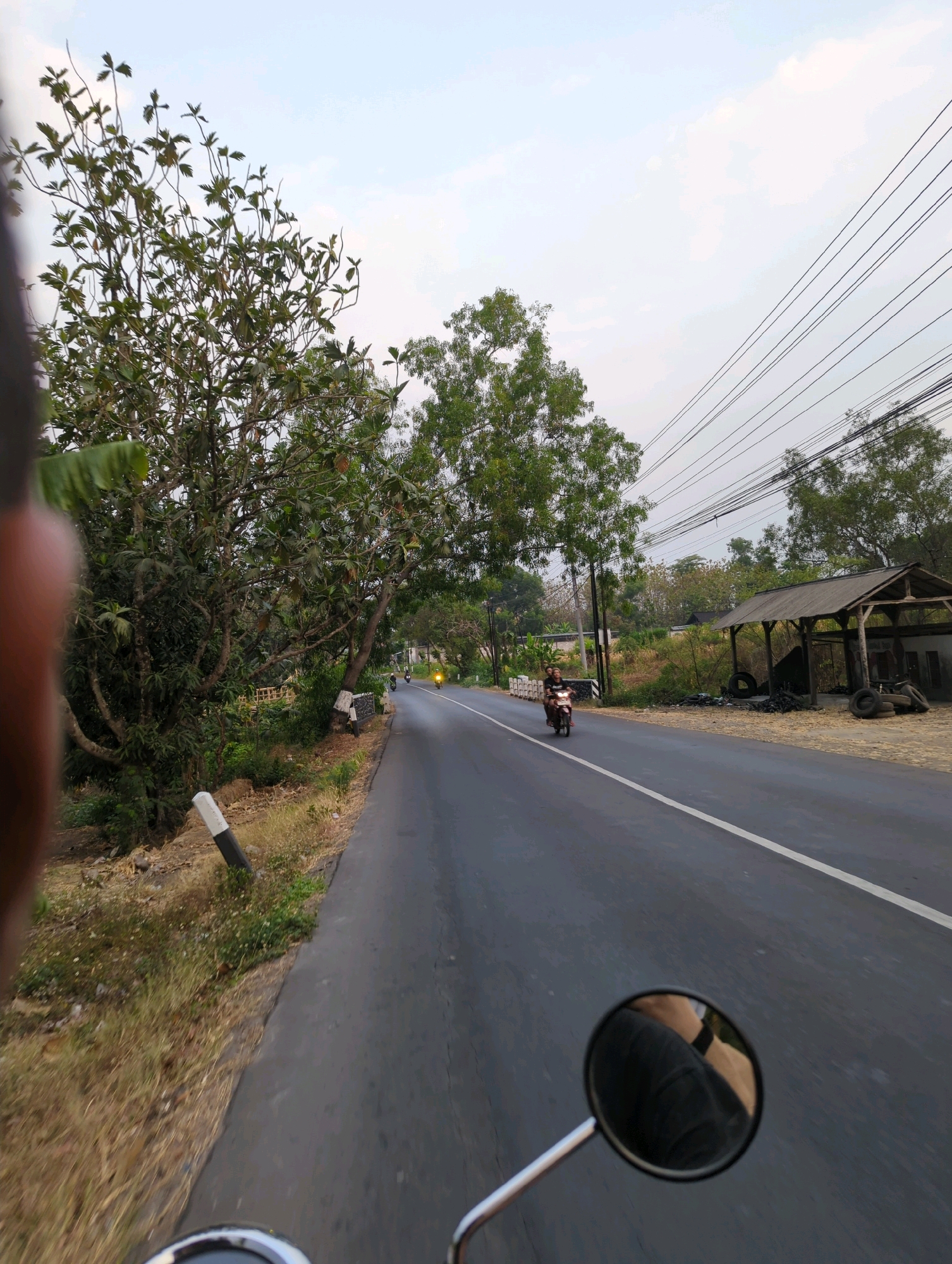 The image depicts a view from a moving vehicle, likely a motorcycle, on a paved road in Senggrong, Indonesia. The road has a white line marking its center. On the left side of the road, there are trees with dense foliage and dry grass. A black and white striped marker post is visible along the roadside. On the right side of the road, there's a rustic open-sided shelter with a thatched roof, stacked tires, and a white building behind it. Several trees are also present on the right side, interspersed with vegetation. Two motorcycles are visible on the road. One, closer to the foreground, carries two people. Another motorcycle, further down the road, has its headlight illuminated, suggesting it is traveling in the same direction as the viewer. The sky is overcast.