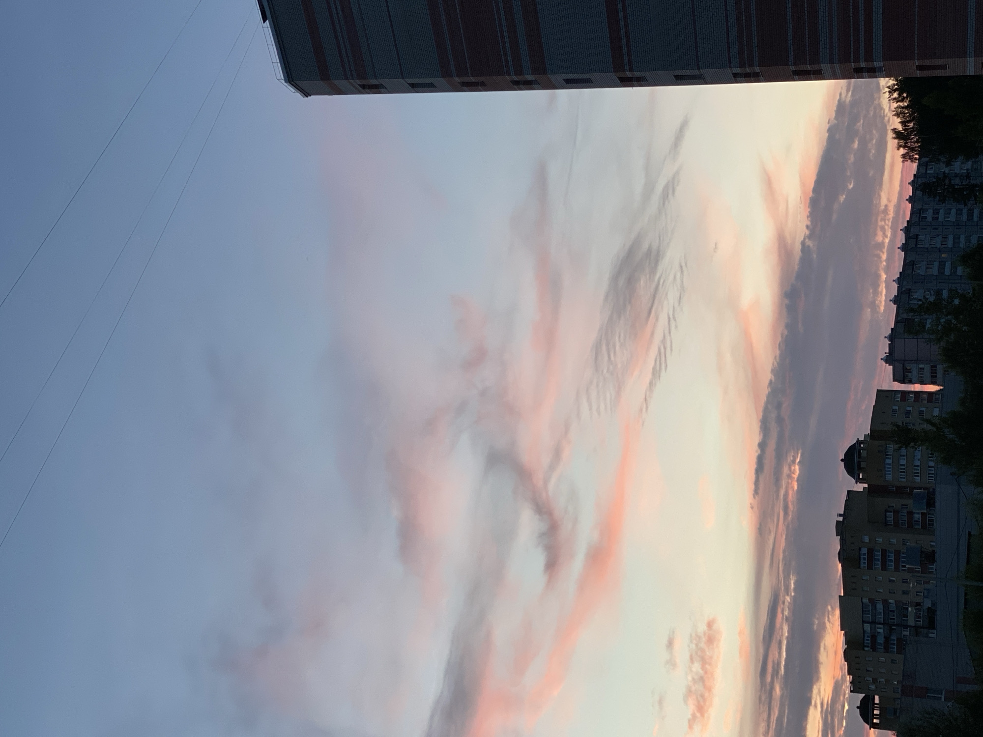 This is an outdoor, vertical, medium-shot photograph of a city skyline at dusk. The sky is a gradient of pale blue at the top, transitioning to hues of pink and orange near the horizon. Wispy, pink-tinged clouds stretch across the sky. In the foreground, on the right side of the frame, is a portion of a tall, multi-story building with alternating bands of dark gray and reddish-brown brick. The left side of the frame shows a row of apartment buildings of varying heights, some with domed architectural features. Trees are visible in the lower portion of the image, obscuring the bases of the apartment buildings. Three thin, dark lines, likely power lines, are visible in the upper portion of the sky. The image was taken in the city of Cherepovets, Russia.