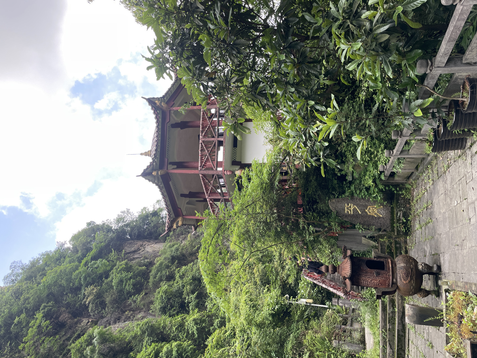 A downward angled medium shot captures a pagoda nestled in lush greenery in Wulingyuan, China. The pagoda features a red and beige construction and a multi-tiered roof. The surrounding environment is densely vegetated with trees and foliage. A stone path leads towards the pagoda, and an ornate, dark-colored incense burner stands to the right of the path. A stone tablet inscribed with golden lettering is behind the incense burner. The sky is visible, featuring a mix of clouds and blue patches.