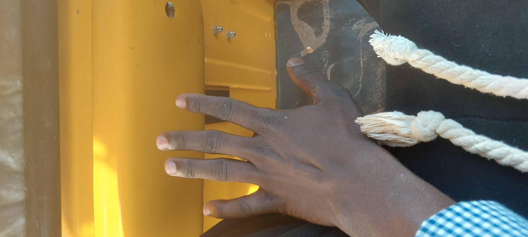 In Jalingo, Nigeria, a person's dark-skinned hand is positioned on a yellow surface, likely the interior of a vehicle. The hand is open, with fingers extended and slightly bent, revealing the fingernails. Below the hand, two thick, light-colored ropes are visible, one with a knot and frayed ends, the other also showing frayed ends. The background consists of a dark, possibly fabric surface.
