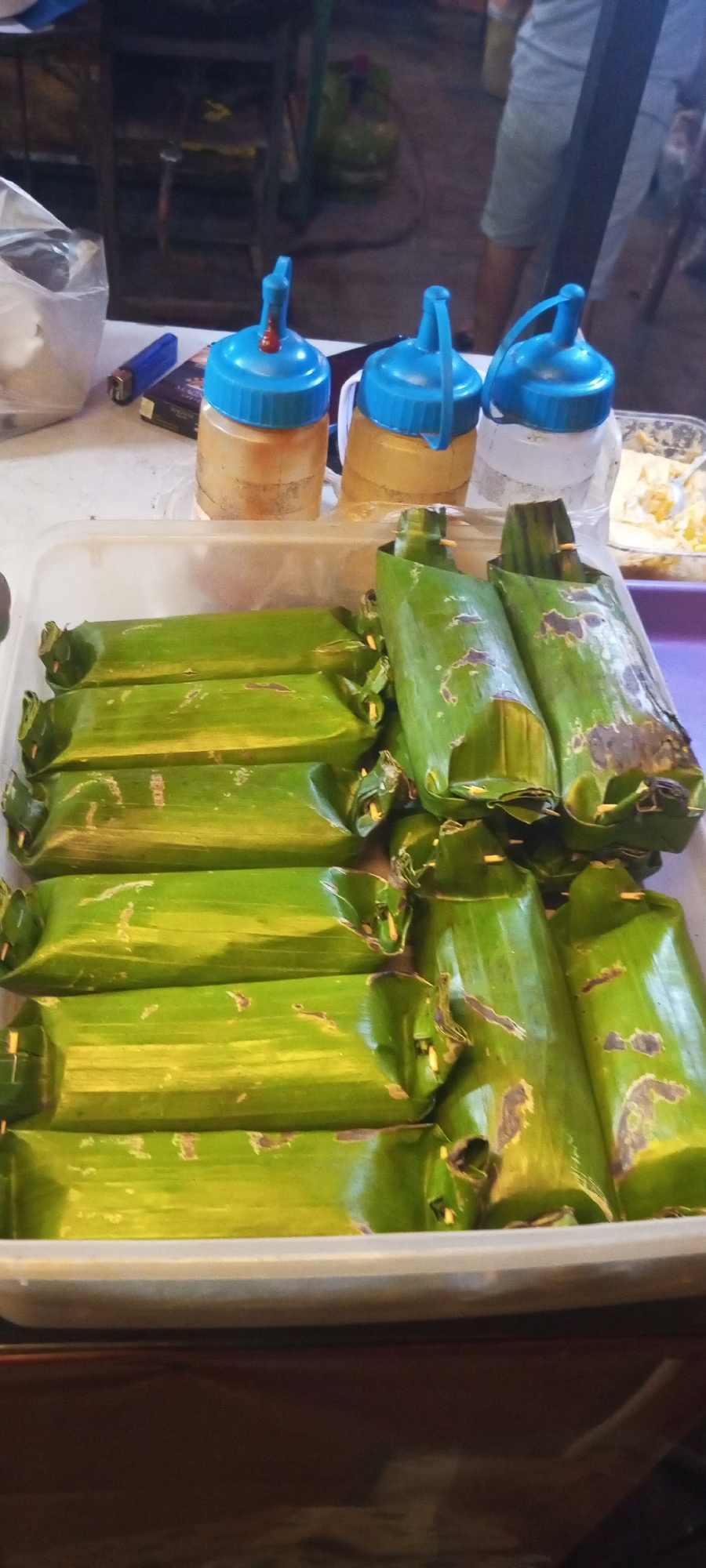 In Setu, Indonesia, a close-up shot captures a white rectangular plastic container filled with several food items wrapped in banana leaves. These food items are cylindrical in shape, with some appearing charred or discolored from cooking. The container is placed on a table, and behind it, three squeeze bottles with blue caps are visible. To the left of the bottles, a blue lighter and a pack of cigarettes are present. The background is blurred, showing what appears to be a market stall with cooking equipment and a person in the distance wearing shorts.