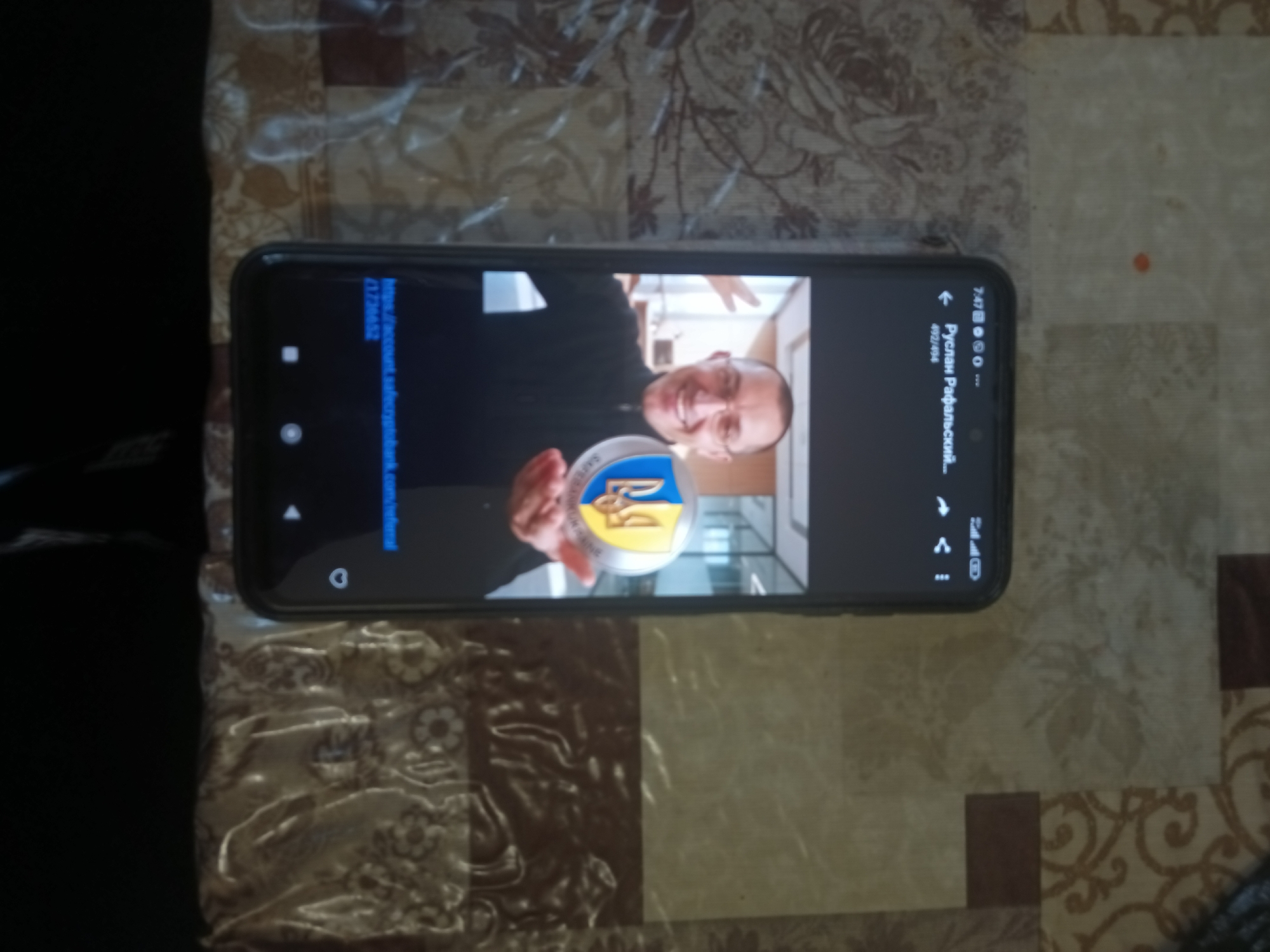 A smartphone is lying on a patterned surface, displaying a video of a man. The man, wearing glasses, smiles and holds up a coin with the Ukrainian trident (Tryzub) emblem. The phone screen shows the timestamp "7:47" and a battery icon indicating a full charge. Below the video, text reads "РусЛан РафаЛьский--" and "492/494". A web address is partially visible on the left side of the screen. The background appears to be a table or countertop with a beige and brown floral pattern. The image is oriented such that the phone is horizontal. The context does not provide any information about the location being in the city of Kyiv, Ukraine.