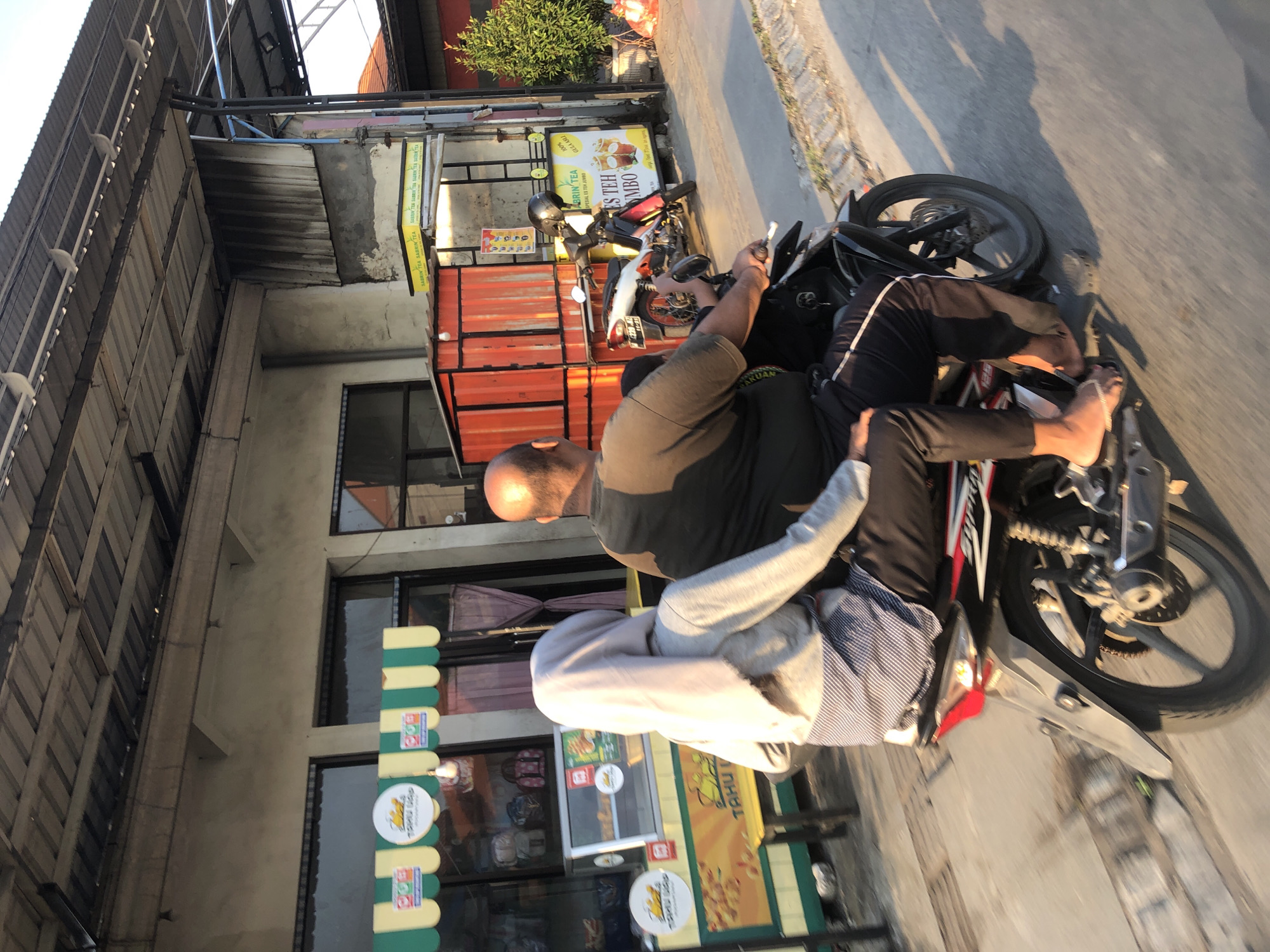 The image depicts a man and a woman riding a motorcycle on a street in Boro, Indonesia. The man, who is driving, is bald and wearing a dark t-shirt and black pants. The woman is wearing a light gray hijab and a gray long-sleeved shirt with a striped pattern on the lower torso. She is seated behind the man, with her arms wrapped around him. The motorcycle is black and red.
In the background, there are several food stalls. To the left, a stall with a green and yellow awning displays pictures of food and a logo that reads "TAHU GEPUK". A glass display case shows food items. To the right, a red stall stands next to a cart with a yellow sign advertising "SABRIN TEA" and "ES TEH JUMBO". A parked motorcycle is visible next to this stall, with a black helmet on its handlebars.
The environment appears to be a commercial area with buildings and paved roads. The lighting suggests it is either early morning or late afternoon, with sunlight casting long shadows. The overall scene is a common sight in a bustling Indonesian city, capturing a moment of everyday life.