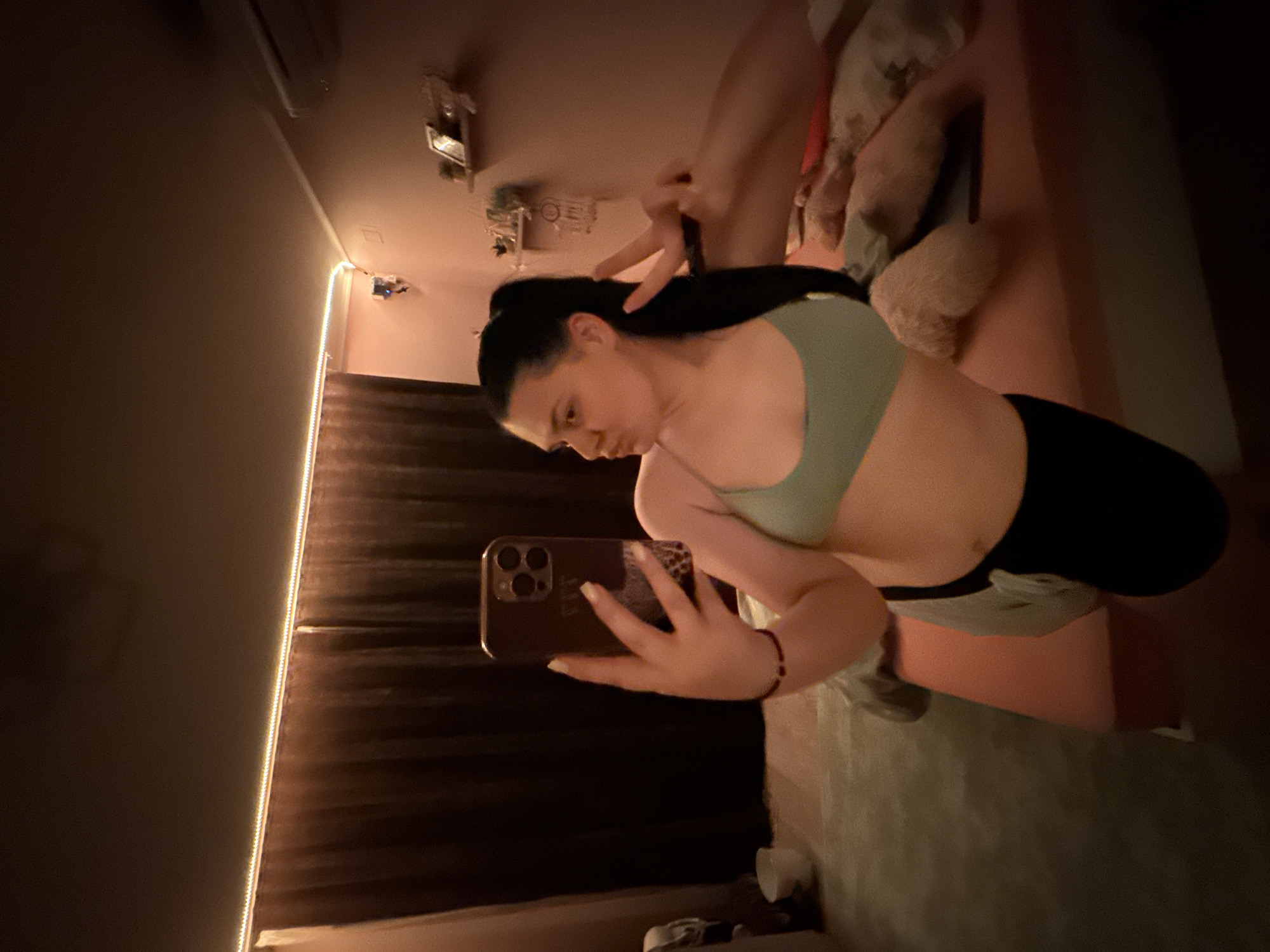 A young woman with dark hair pulled into a ponytail takes a selfie in a dimly lit bedroom. She is wearing a light green sports bra and black shorts. Her left arm is extended, holding a smartphone, while her right hand is raised, making a peace sign. The room appears to be a bedroom with a bed visible in the background, covered in light-colored bedding and pillows. Dark curtains hang on one side of the room, and LED strip lights are illuminating the ceiling. The overall ambiance is warm and intimate, with soft lighting casting shadows. The location is specified as Rogosh, Bulgaria.