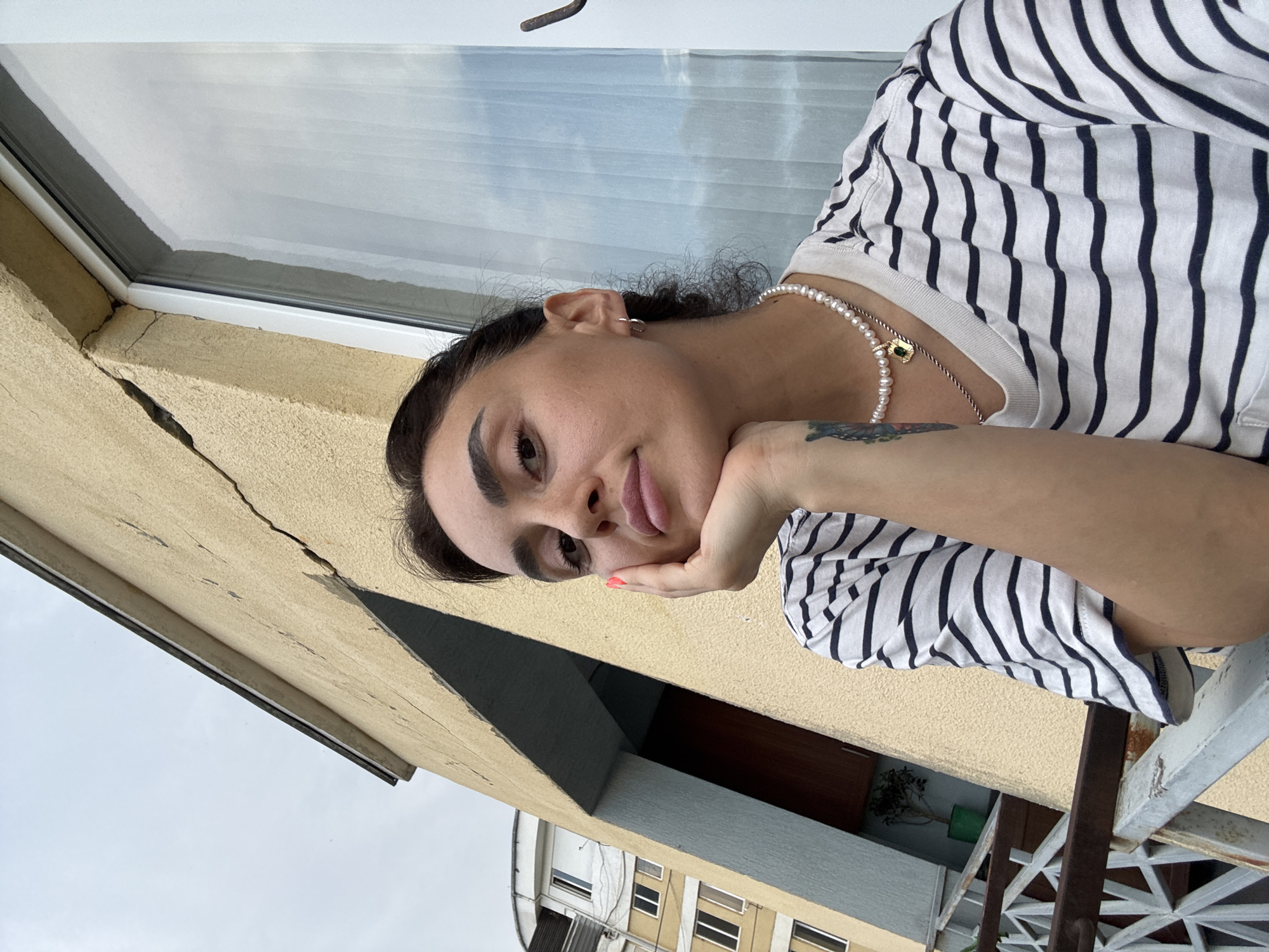 A woman is taking a selfie on a balcony in Tbilisi, Georgia, with a building and the sky in the background. She is wearing a striped t-shirt and a pearl necklace. Her hand is resting on her chin. The building behind her is yellow with a large crack in the wall. To the left, another apartment building is visible. The sky is overcast.