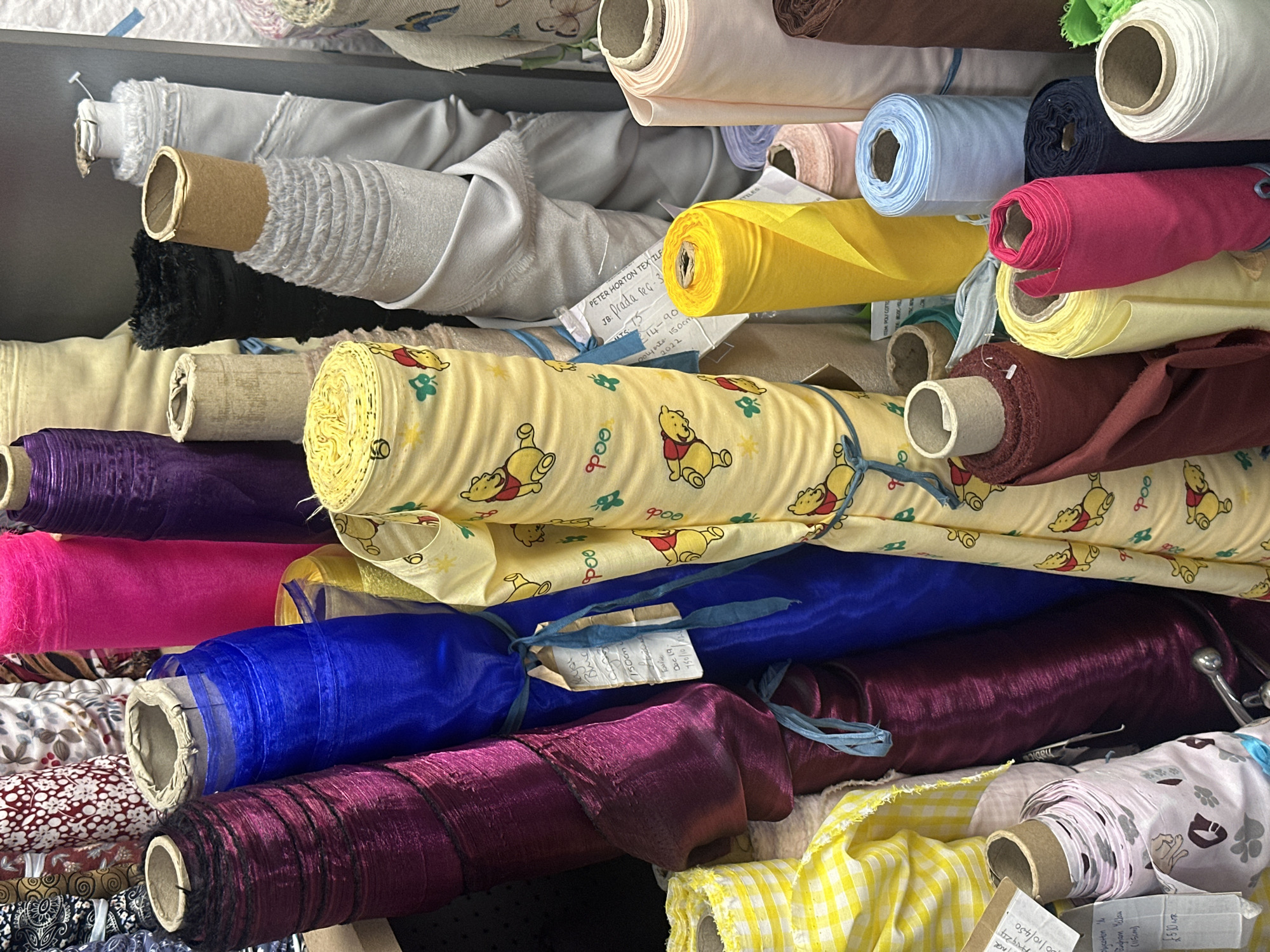 A densely packed collection of fabric rolls fills the frame. The fabrics vary in color, pattern, and texture, creating a visually rich tableau. Prominent colors include yellow, blue, purple, pink, and white. Several rolls feature patterns, such as Winnie-the-Pooh characters, floral designs, and checks. The fabric rolls are stacked haphazardly, with some labels and tags visible. The rolls appear to be in a warehouse. There is no evidence of the image being located in the city of Sevenoaks, United Kingdom.