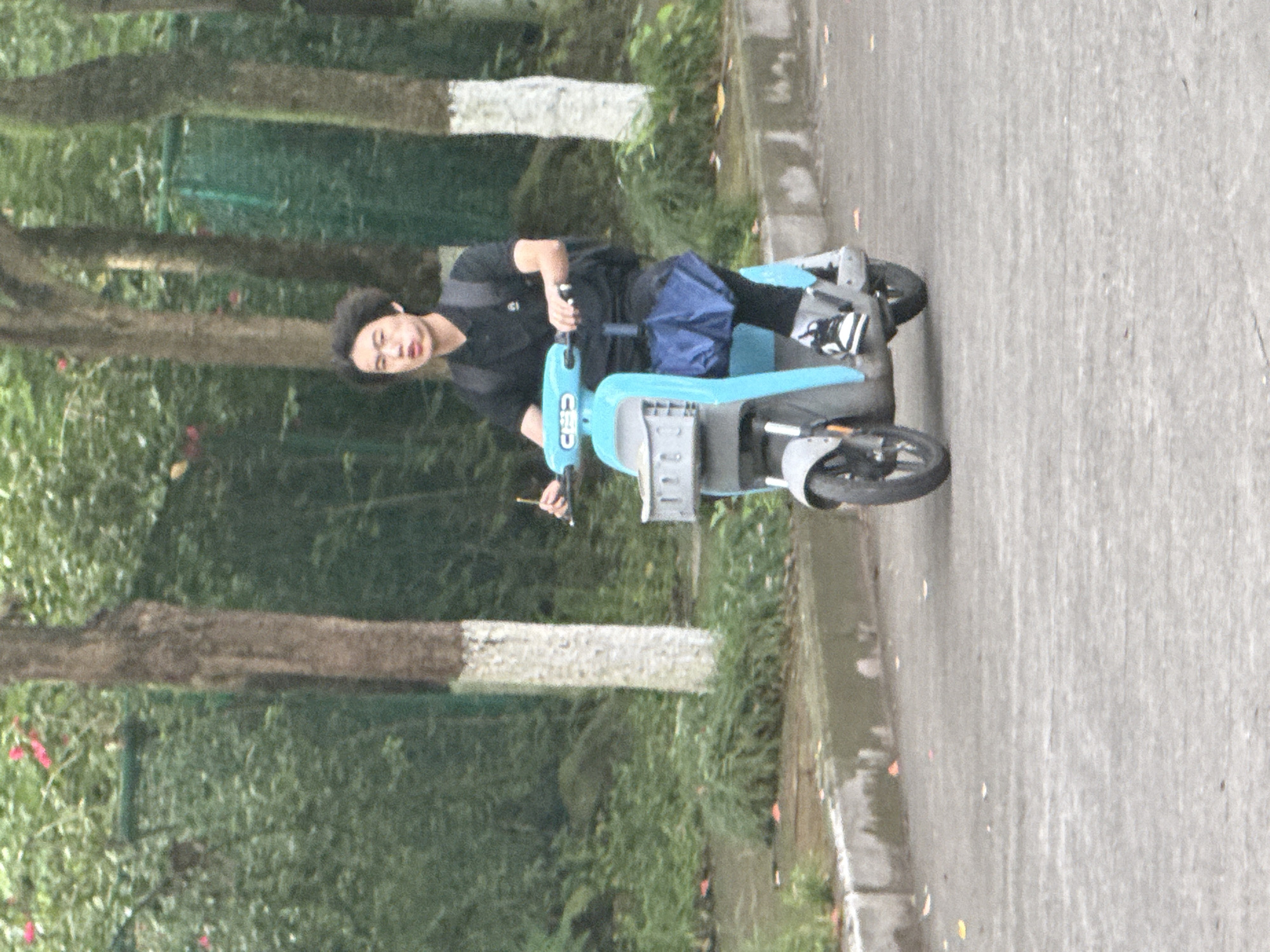 A person is riding a light blue electric scooter on a wet paved road. The person is wearing a black shirt, black pants, and black and white sneakers. They have short, dark hair and are wearing a beret. They are looking to their left. The scooter has a small front wheel and a larger rear wheel. A blue bag is on the scooter. There is green foliage on the left side of the frame, including trees with white-painted sections. The road is wet with rain. The location is Jiangjin, China.