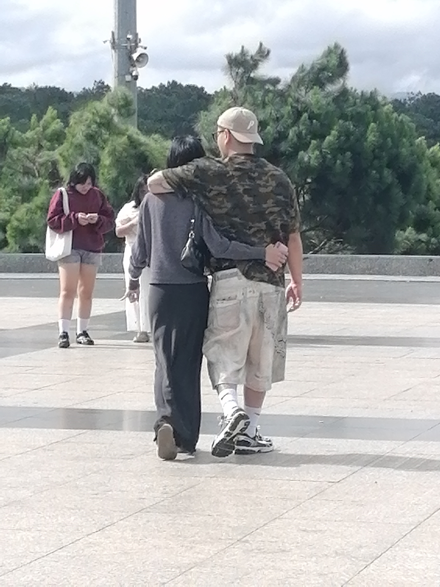 In Da Lat, Vietnam, a man wearing a camouflage t-shirt, light-colored shorts, and a beige baseball cap with a "P" logo is seen from behind, with his left arm around a woman. The woman, dressed in a dark gray long-sleeved shirt and a long dark skirt, is also viewed from behind, with her left arm around the man's waist. They are walking away from the camera on a paved outdoor area. To their left and further in the background, a woman in a maroon hoodie and shorts, carrying a white tote bag, looks down at her phone. Behind her, another person in a light-colored outfit is partially visible. The environment includes lush green trees and a cloudy sky, suggesting an overcast day.
