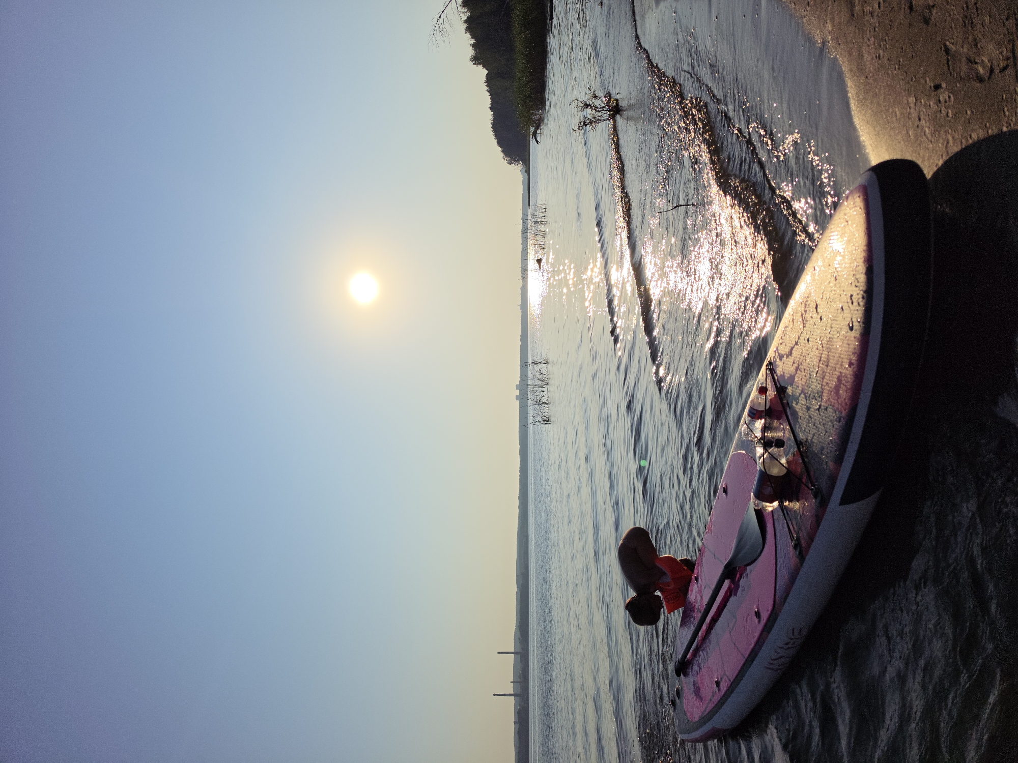 In Chaykovsky, Russia, an individual wearing an orange life vest is partially submerged in the water next to a pink and white stand-up paddleboard. The paddleboard is equipped with a paddle and two plastic bottles, one of which appears to be a water bottle. The scene unfolds at the edge of a body of water, with gentle waves lapping at a sandy shore. In the distance, across the water, industrial structures including smokestacks are visible, along with a treeline and reeds. The sun, low in the clear sky, casts a bright reflection on the water's surface, creating a shimmering effect.