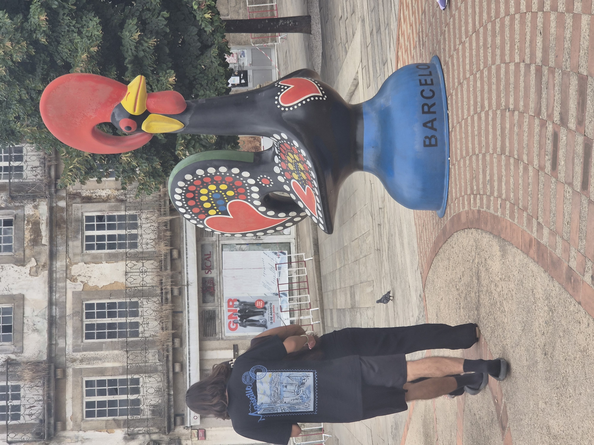 The image depicts a public square in Barcelos, Portugal, dominated by a large, black, decorative rooster statue with a red comb and wattle, yellow beak, and colorful patterns of hearts and dots. The statue stands on a blue base inscribed with the word "BARCELOS." In the background, an old building with weathered facades and balconies with ornate iron railings is visible, along with trees and what appears to be a shopfront with posters. Two individuals, one wearing a black t-shirt with a graphic of Marseille and the other in dark clothing, are walking away from the camera. A pigeon is on the cobblestone ground in front of them. The ground is paved with a pattern of brick and stone.