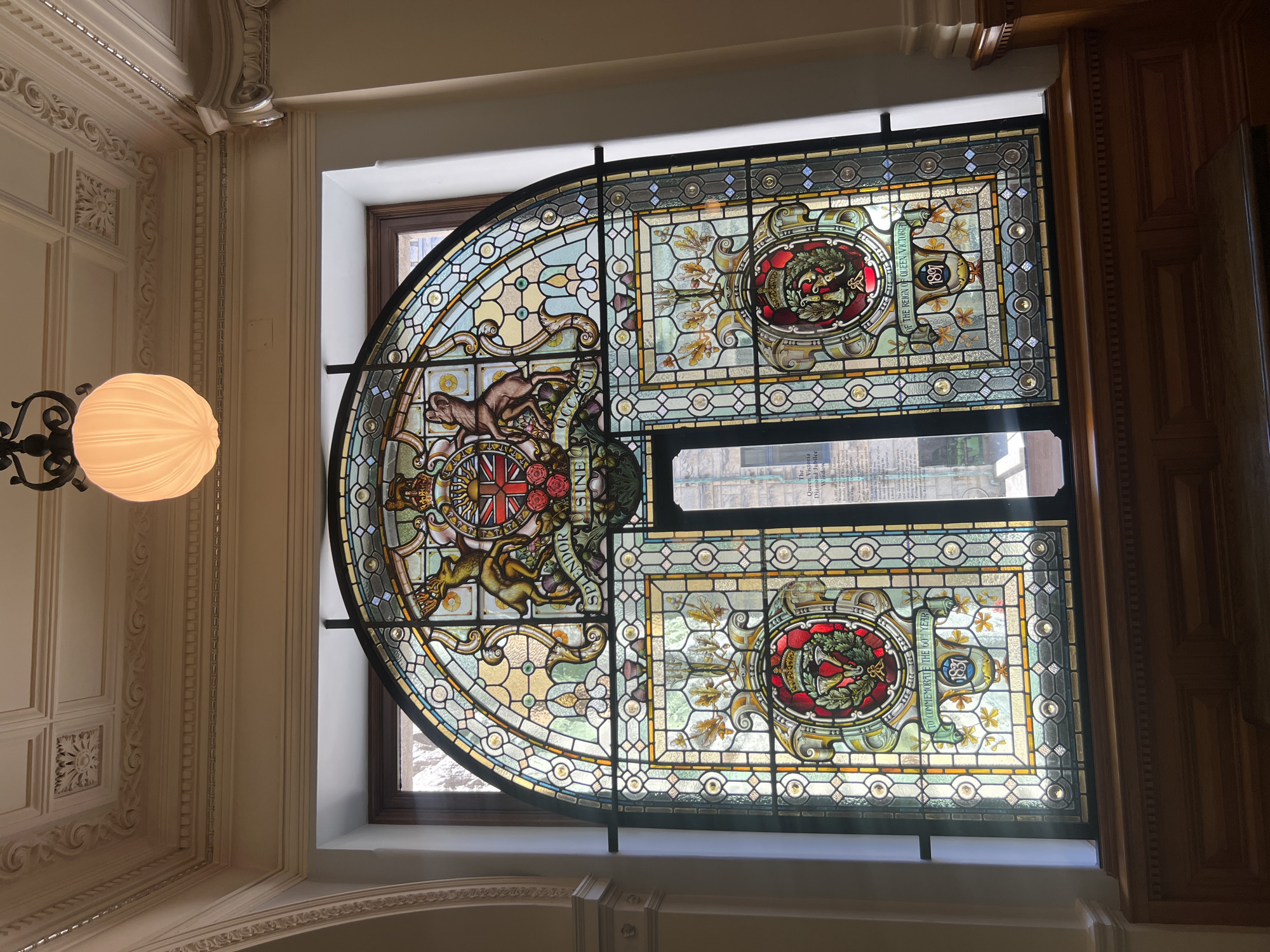 The image shows a large, ornate stained-glass window, likely in a historic building. The window is arched at the top and divided into several sections. The central top portion features a coat of arms with a lion and a unicorn, along with the Latin phrase "SPLENDOR SINE OCCASU." Below this, on the left and right sides, are circular emblems each containing a crown and a floral design. The bottom section includes the inscription "TO COMMEMORATE THE 60TH YEAR" and "OF THE REIGN OF QUEEN VICTORIA 1897". The window is set within a wooden frame and is illuminated by natural light filtering through it, casting colored patterns on the interior. A decorative light fixture hangs from the ceiling above the window. The overall impression is one of grandeur and historical significance. This window is located in the Legislative Assembly of British Columbia in Victoria, Canada, commemorating Queen Victoria's Diamond Jubilee.