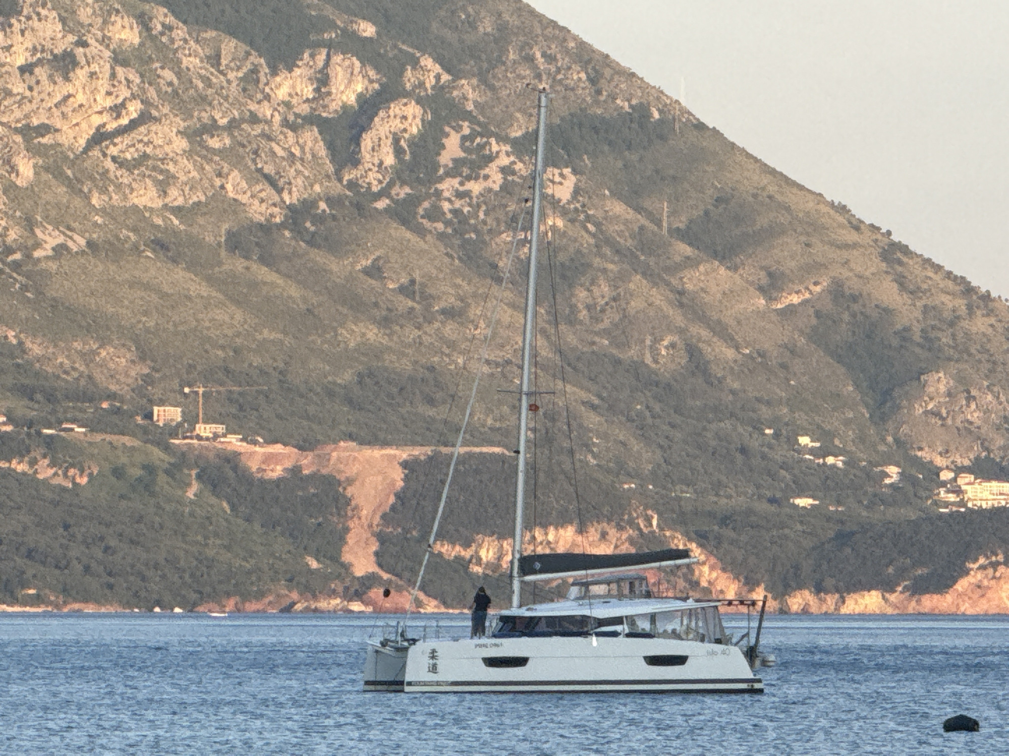 A white catamaran, bearing the name "Iulo 40" and Japanese characters on the hull, is anchored in calm blue water. A person stands on the bow of the boat. In the background, a large mountain dominates the landscape. A construction crane is visible on the mountain slope. The image was likely taken near Budva, Montenegro, based on the coastal terrain and architectural style.