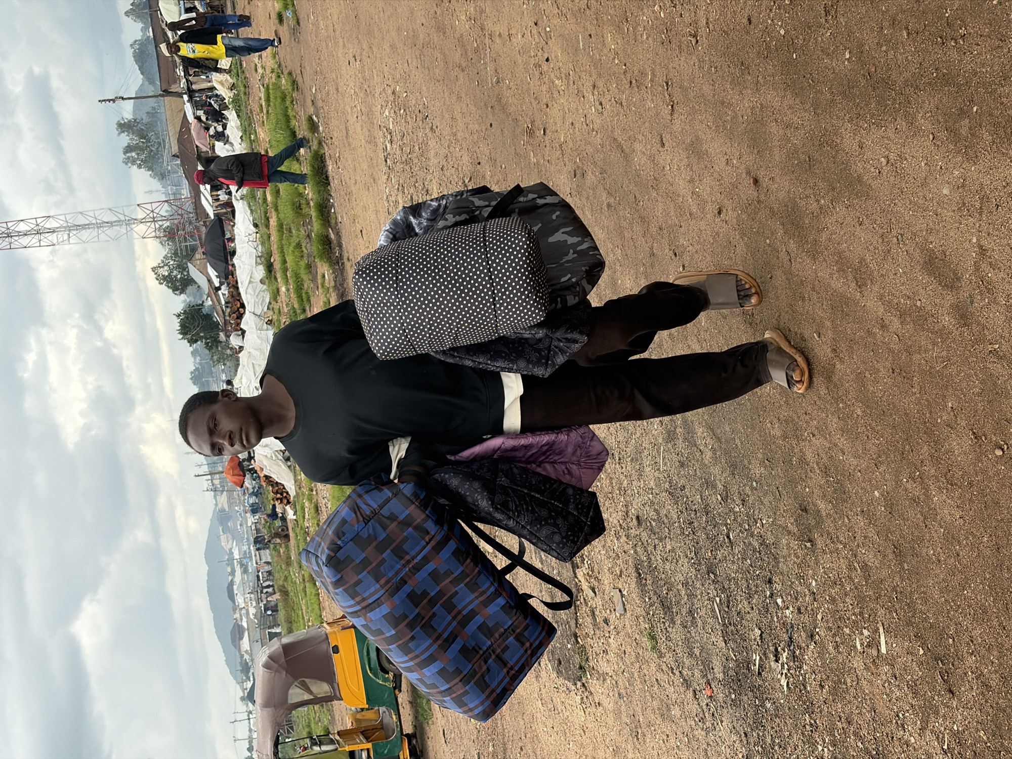 A young man walks on a dirt road in Bukuru, Nigeria, carrying several large bags. He is wearing a black t-shirt and dark pants, with a pair of open-toed sandals. In his arms, he holds a large, checkered blue and black duffel bag, a smaller black patterned bag, a purple folded item, and a large white cylindrical bag with black polka dots. To his left, a yellow and green auto-rickshaw is parked. In the background, a market scene unfolds with various stalls, people, and a tall radio tower. The sky is overcast, and the ground is dry and dusty.