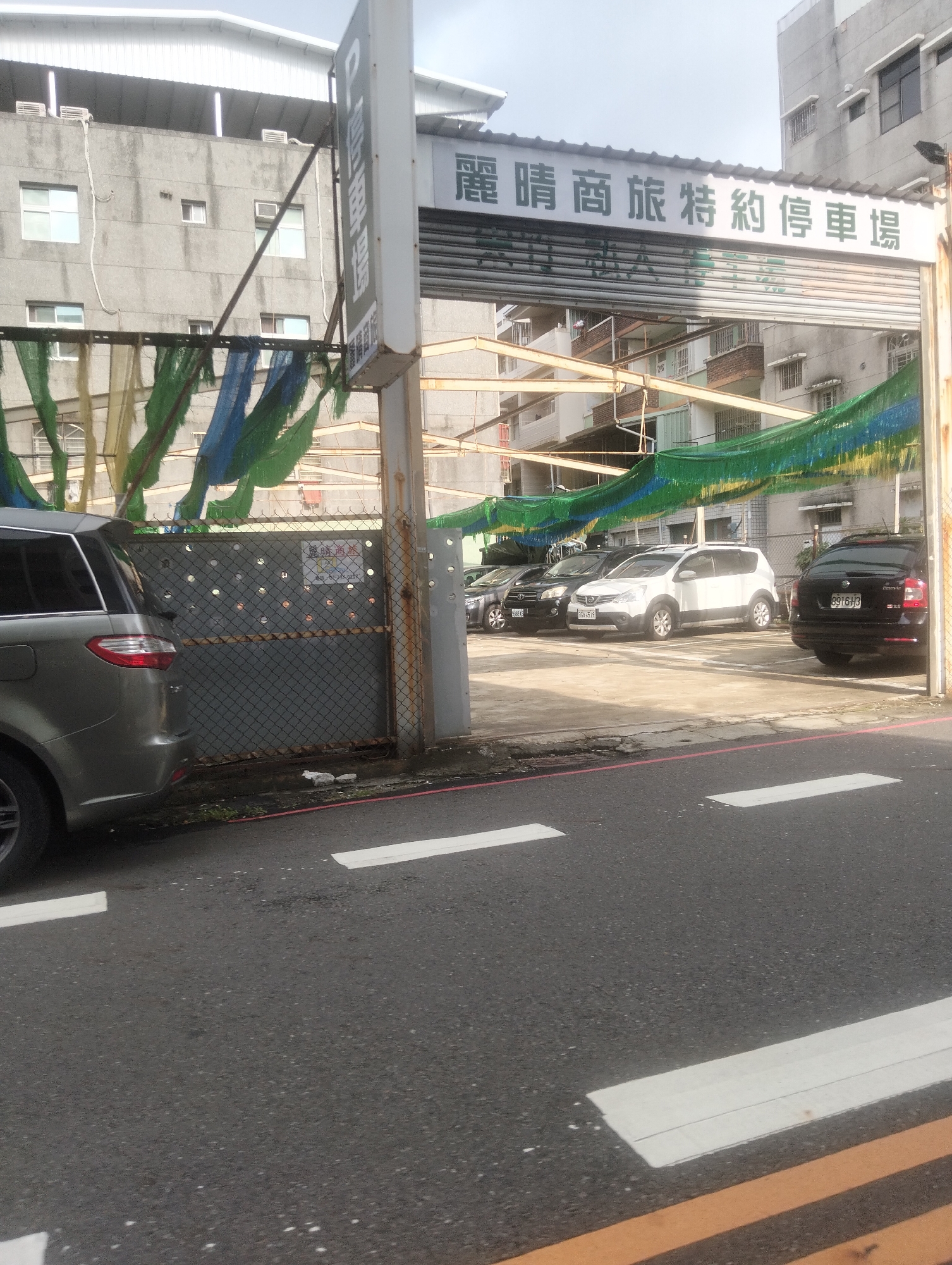 A grey SUV is parked in front of a chain-link fence with a patterned design, partially obscured by a green and blue tarp. Beyond the fence, several cars are parked in a lot, including a white SUV, a black sedan, and a black station wagon. A tall, white sign with Chinese characters stands to the left of the fence, topped with a metal structure supporting the tarp. The sign is vertically oriented and features a white rectangle with dark text. Above the parking lot, a large white awning extends over the entrance to the lot, displaying more Chinese characters. The building behind the lot is a multi-story concrete structure with visible windows. The foreground shows a dark asphalt road with white lane markings. The scene appears to be a parking area in an urban environment, likely in Kaohsiung, Taiwan, given the context of the Chinese characters on the signage.