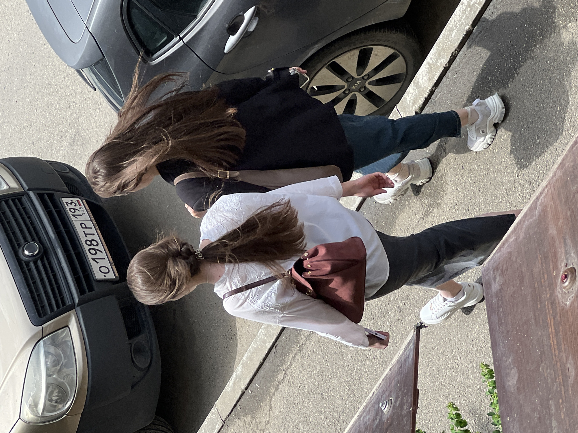 Two women are walking on a sidewalk next to parked cars. The woman in the foreground wears a white blouse, black pants, and white sneakers, and carries a burgundy shoulder bag. The woman behind her wears a black jacket, blue jeans, and white sneakers. A black car and a beige van with license plate "O198TP 193 RUS" are parked nearby. The setting is an urban environment, likely Krasnodar, Russia, given the license plate. A wooden bench and some greenery are visible on the right side of the frame.