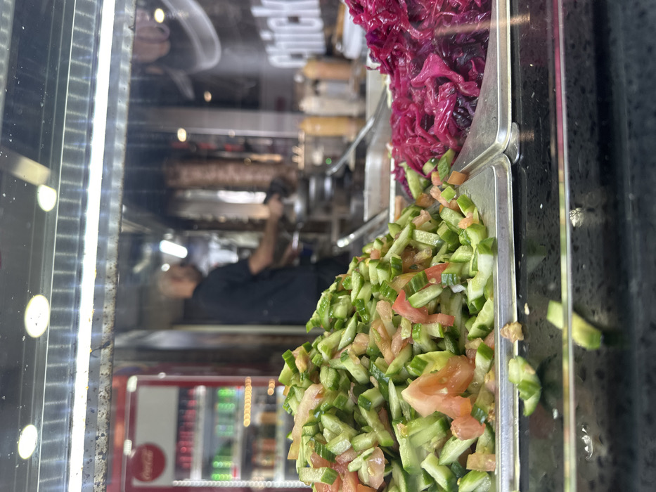 The image displays an interior view of a food service establishment in Mishmar Hasharon, Israel, observed through a transparent barrier. In the foreground, two metal trays rest on a dark, reflective counter. The left tray contains a large mound of diced cucumbers and tomatoes. The right tray holds a pile of shredded, purple-red pickled cabbage. Scattered pieces of cucumber are visible on the dark counter directly below the trays.
In the midground, behind the transparent barrier, a blurred male individual with a bald head, wearing a dark shirt, is visible. He stands behind a counter, facing away from the camera, and is engaged in an action related to food preparation, with his right hand near a large, vertical rotisserie skewer of cooked meat (shawarma/doner kebab). To the left of the individual, a red Coca-Cola branded refrigerator containing bottled beverages is present. To his right, several squeeze bottles, likely containing sauces, are visible on the counter, along with a partially visible sign displaying the word "Chick." Overhead, fluorescent light fixtures illuminate the scene.