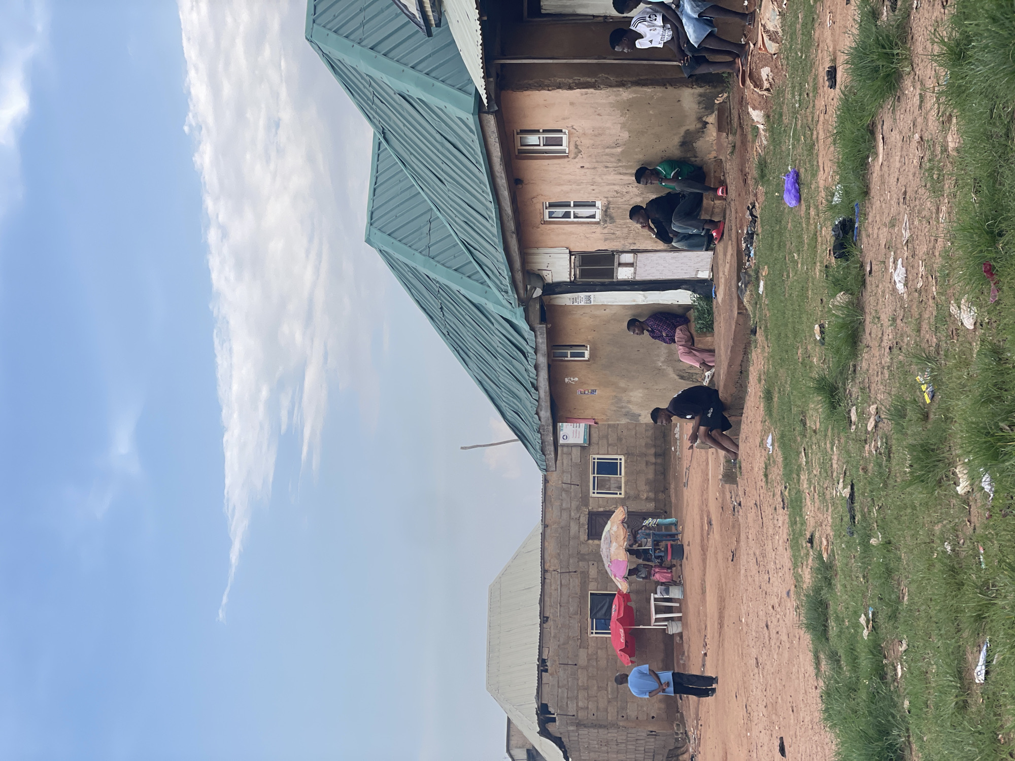 In Kurunduma, Nigeria, a scene unfolds with several individuals gathered outside a row of buildings under a cloudy sky. In the foreground, the ground is a mix of red dirt and sparse, patchy green grass littered with debris. To the right, a young man in a black t-shirt and shorts sits on a concrete ledge, looking down. Further back, a group of young men are seated on the ground or ledges along the front of a textured, earth-toned building with a green metal roof. One man wears a plaid shirt and light pink pants, another a black t-shirt and jeans, and a third a green t-shirt and jeans. To the far right, two more young men are seated, one wearing a white and black sports jersey with the logo "Emirates" and blue shorts, and another wearing a black t-shirt and blue shorts.
To the left of the earth-toned building, another building constructed of cinder blocks is visible. In front of this building, a man in a blue shirt and dark pants stands facing away from the camera. Near him are two outdoor umbrellas, one red and one colorful, sheltering a white table with a bucket. A small group of people, including women and children, are standing or sitting near the table. The overall environment suggests a residential or commercial area, possibly a market or a gathering spot, with signs of everyday life in a Nigerian community.