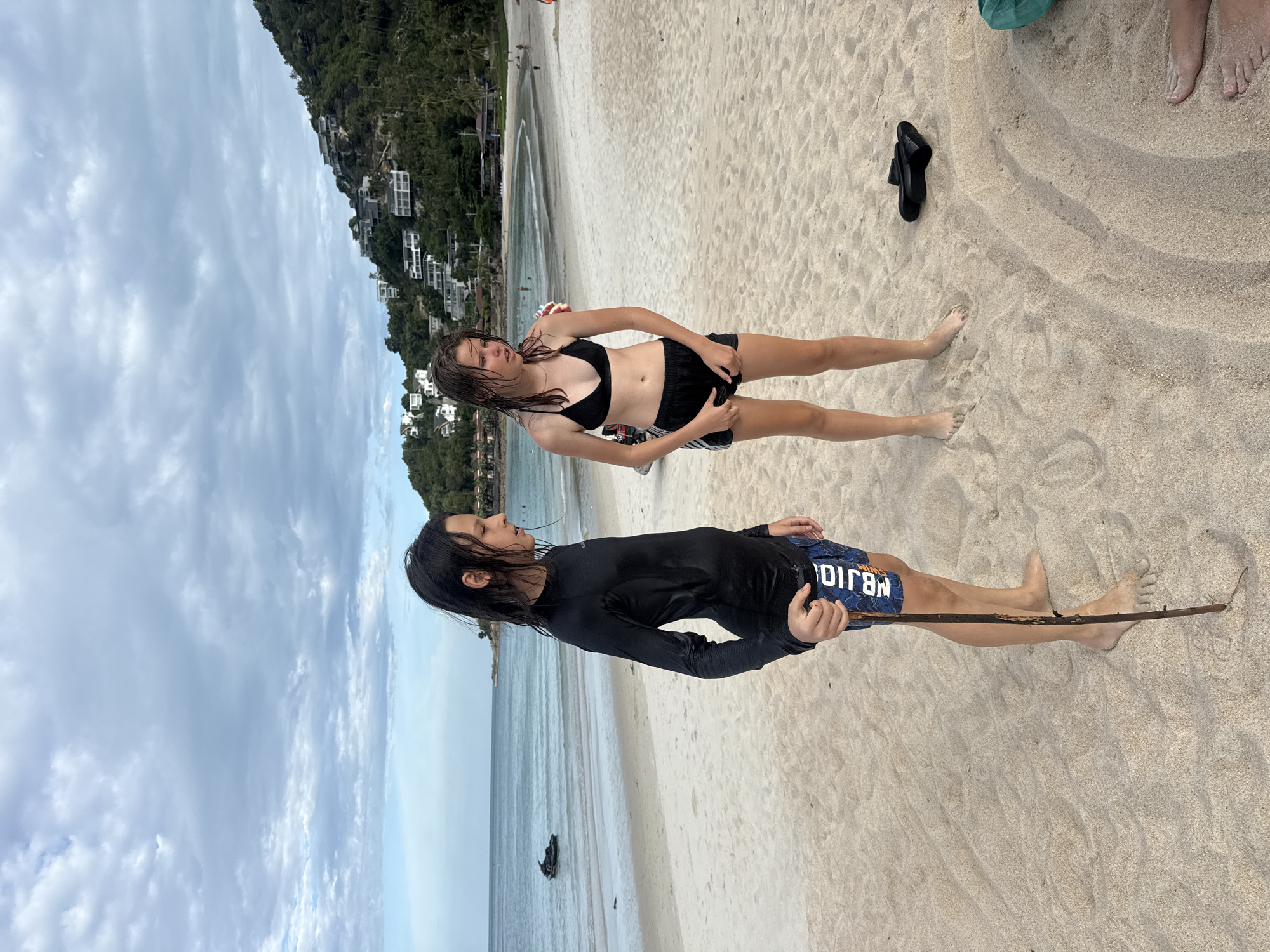 The image depicts a beach scene in the Ko Samui District of Thailand. In the foreground, two young individuals stand on a sandy beach. The person on the left, with long dark hair, is wearing a black long-sleeved rash guard and patterned swim trunks. They are holding a long stick. The person on the right, with light brown hair, is wearing a black bikini and black shorts. The background shows the ocean with a jet ski in the distance, and a hillside covered in lush green vegetation with buildings nestled into it. The sky is overcast with dramatic clouds.