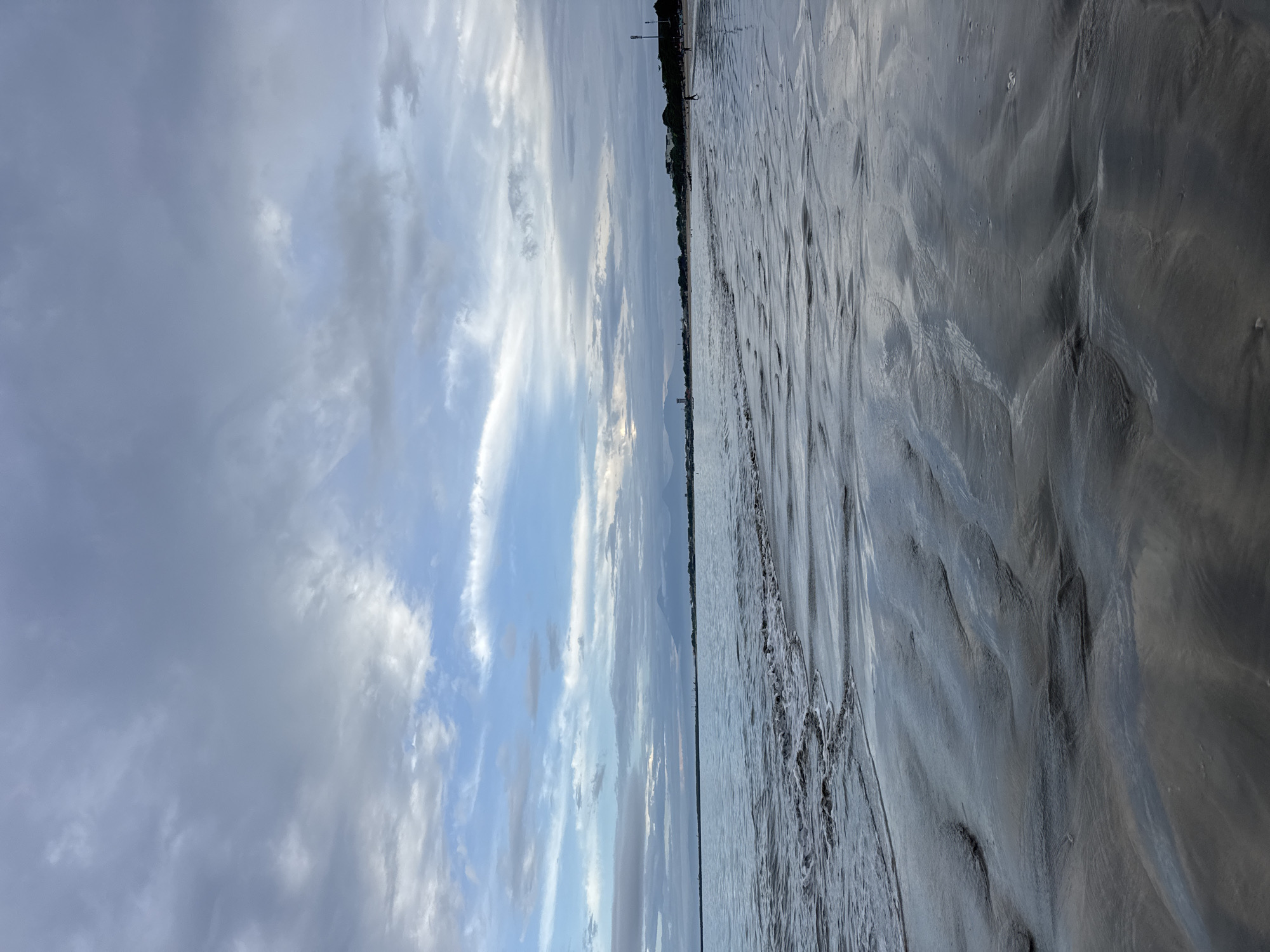 The image, taken in Kuta Selatan, Indonesia, displays a vast, open landscape composed primarily of a cloudy sky and a textured wet sandy or muddy ground. The scene is depicted from a low vantage point, looking across the expanse.
The upper portion of the image is dominated by a dynamic sky filled with a mix of cirrus and stratocumulus clouds. Patches of brighter blue sky are visible between the clouds, particularly towards the center and right, suggesting either partial clearing or the subtle glow of the sun, possibly during early morning or late afternoon. The clouds vary in tone from light gray and white to darker, denser formations, creating a sense of depth and movement.
A faint horizon line stretches across the mid-ground, separating the sky from the ground below. Along this distant horizon, a very subtle, dark outline of land or a coastline is discernible, featuring what appears to be a few distant structures or sparse vegetation as dark silhouettes.
The lower and larger part of the image showcases an intricate pattern of wet sand or mud, possibly at a beach or tidal flat. The surface is highly textured, displaying numerous ripples, undulations, and channels, likely carved by receding water or tidal movements. These patterns create a visually rich, almost abstract foreground with varying shades of dark gray and silver, reflecting the light from the sky above. No specific human activity or event is visible, and the scene appears natural and untouched. There is no visible text in the image.