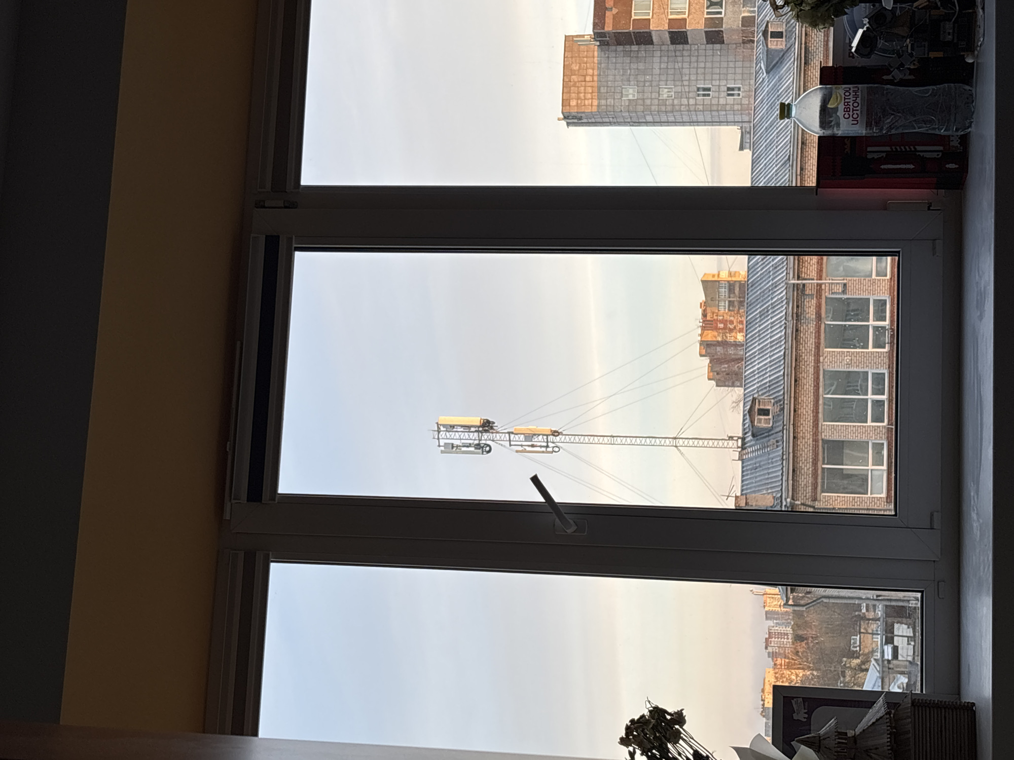 The image shows a view from a window, revealing a cityscape in Perm, Russia. A construction crane is visible against a backdrop of buildings with varied architectural styles, including brick and gray-roofed structures. Items such as a water bottle and various objects clutter the windowsill. The window is partially open, with its handle visible. The sky appears overcast, casting a soft light over the scene.
