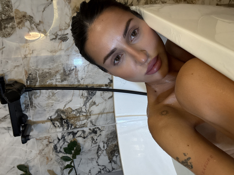 A woman with dark hair and tanned skin lies partially submerged in a white bathtub. She looks directly at the camera. Part of her shoulder and arm are visible, revealing two small tattoos. The bathroom has marble walls. A black adjustable phone holder is attached to the wall to the left. A small green plant sits at the bottom of the frame. The woman's head rests near the tub's edge, angled slightly.