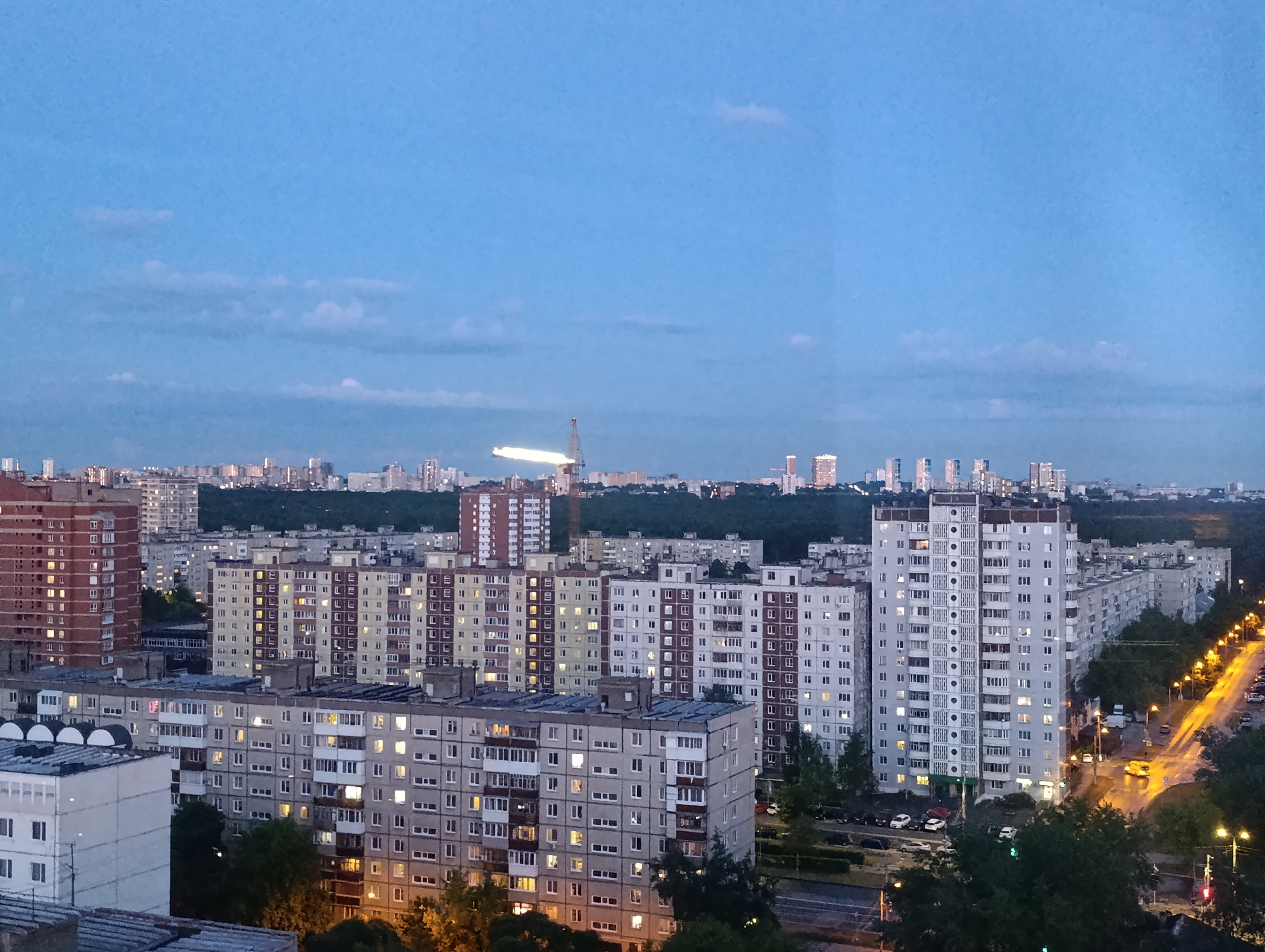 This is an elevated view of a residential urban landscape in Perm, Russia, captured during the twilight hours. The scene is dominated by numerous multi-story apartment buildings, characteristic of post-Soviet architecture, with many windows lit from within, suggesting human activity. The sky above is a deep, clear blue, with a few faint clouds.
In the foreground and mid-ground, a dense cluster of residential blocks fills the frame. These buildings vary in color from reddish-brown to light grey and beige. On the right side of the image, a curving street is visible, illuminated by a series of warm yellow streetlights, under which several cars are parked. Trees and dark foliage line the street and are interspersed among the buildings.
In the far distance, a city skyline with taller, more modern structures can be seen, separated from the immediate residential area by a dark strip of what appears to be a forest or parkland. A prominent, brightly illuminated, elongated structure or object, possibly a crane or a construction site element, stands out against the distant skyline. No specific human activity is discernible, but the illuminated windows and streetlights convey a sense of evening life. The overall atmosphere is quiet and still.
The OCR detected fragmented and illegible text, such as "11", "LE", "E12", "19", "川", "Π", "116", and "101", which likely correspond to indistinct shapes or features on buildings rather than decipherable text.
