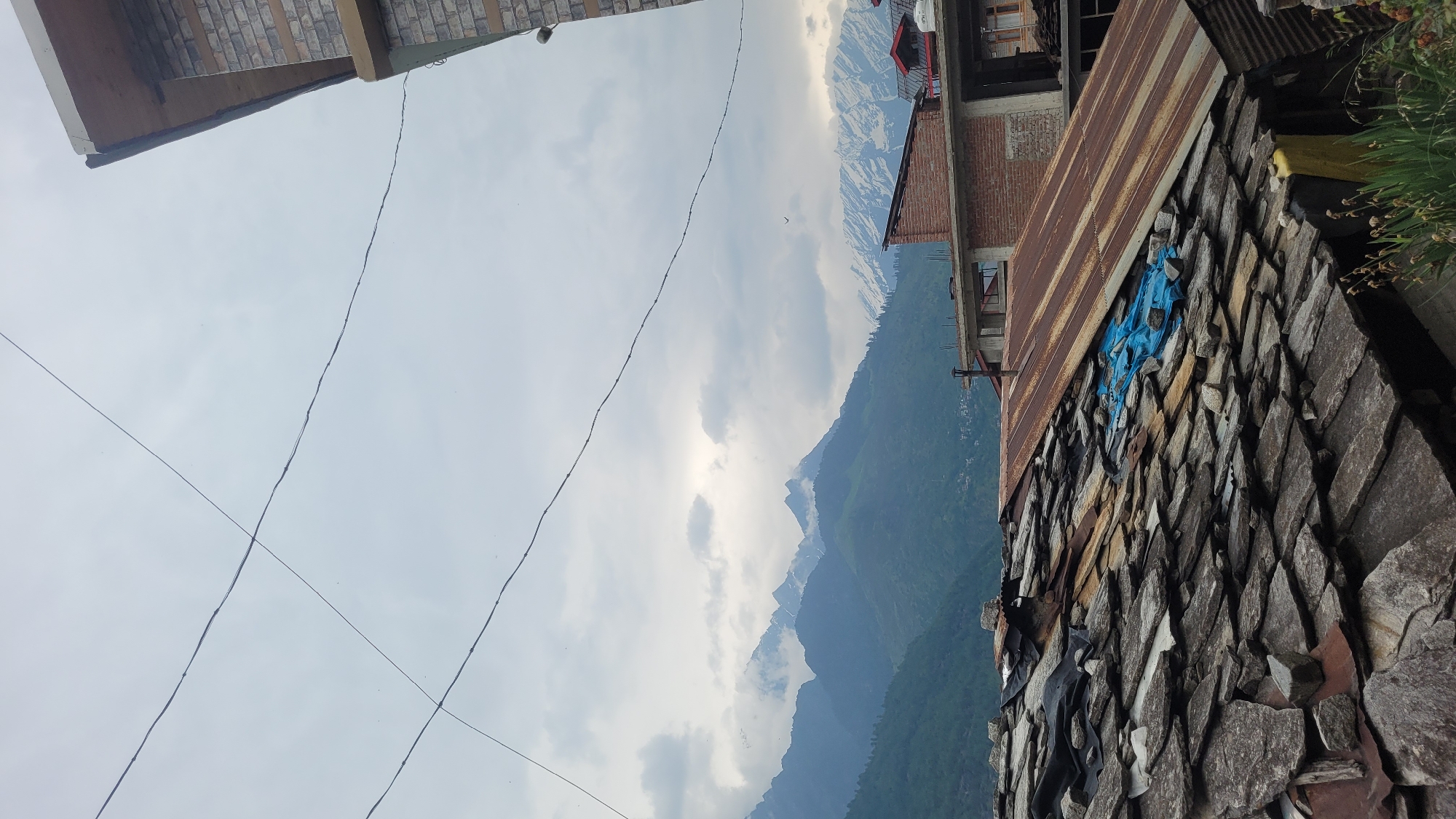 This is a vertical, eye-level shot of a mountainous landscape in Vashisht, India, featuring a cloudy sky, snow-capped mountains, and several structures. In the foreground, a stone-tiled roof is prominent, with a section covered by a rusty corrugated metal sheet and a blue tarp. Behind this, several buildings made of brick and stone are visible, some with balconies and windows. The middle ground is dominated by rolling green hills dotted with trees, leading to a range of majestic, jagged mountains, some of which are covered in snow. Power lines crisscross the cloudy sky. The overall scene conveys a sense of rural, mountainous life in a cloudy weather.