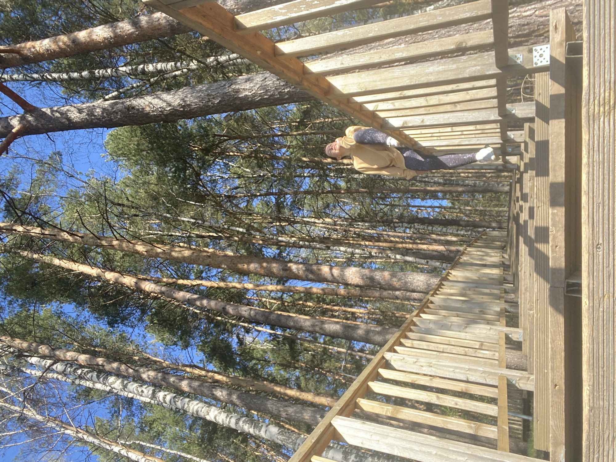 A low-angle shot of a woman lying on a wooden structure within a forest, with the perspective making it appear as though she is suspended in the air. The structure is a wooden walkway or bridge, likely elevated within the trees. The woman is wearing a light brown jacket and patterned leggings. The trees are tall and dense, obscuring the sky. The photo appears to have been taken in Chaikovsky, Russia, based on the metadata.