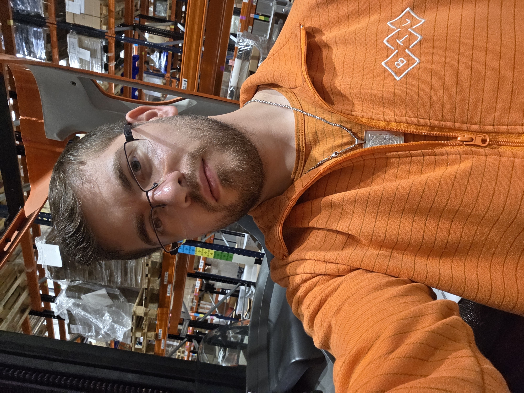 A man with brown hair and a beard is wearing glasses and an orange zippered hooded sweatshirt with an embroidered logo. He is inside a warehouse with shelves stocked with various packages. The location is likely in an industrial area of Plovdiv, Bulgaria, due to the presence of logistics and distribution centers in the city.