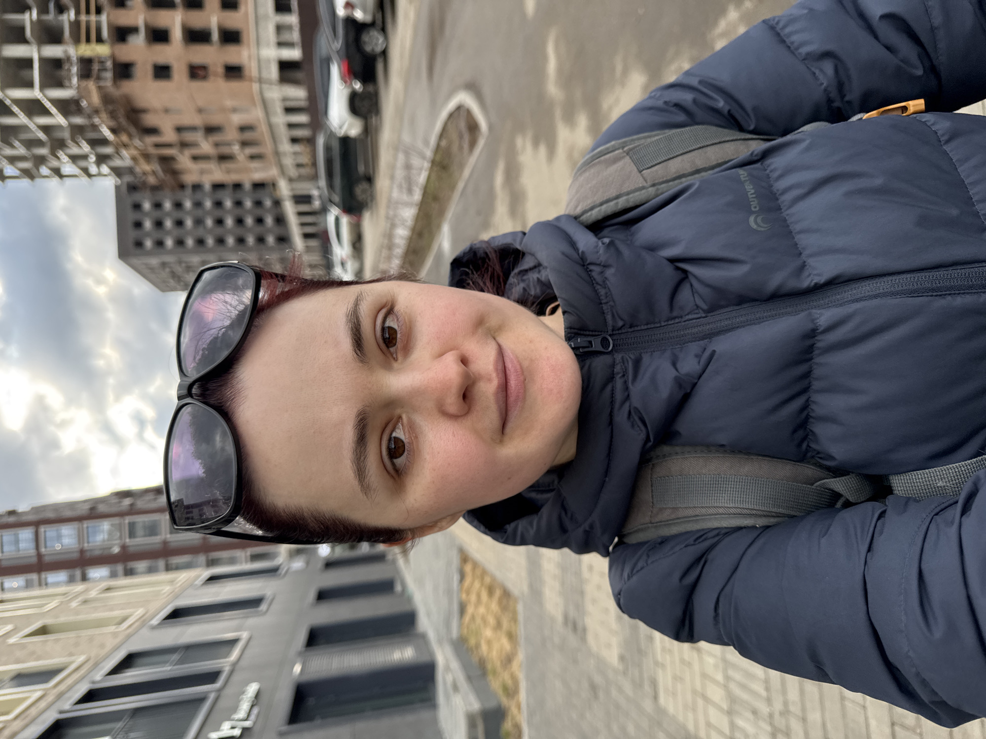 A woman is captured in a selfie-style shot, looking directly at the camera with a calm, slight smile. She has light skin, dark brown eyes, and dark reddish-brown hair, with sunglasses pushed up onto her forehead. She is wearing a dark blue puffer jacket, and a grey backpack strap is visible over her right shoulder. A logo, possibly "Outventure," is visible on the jacket's left sleeve.
The setting is an urban outdoor environment under an overcast sky. In the background, to the woman's left, are multi-story buildings, some appearing to be under construction with visible concrete frames and others with windows. To her right, a paved street or pathway features parked cars, including white and dark sedans or SUVs. The ground is paved, possibly with light-colored bricks or patterned concrete. Reflections in the woman's sunglasses show what appears to be trees and parts of buildings or the sky. The time of day is daytime, with muted lighting typical of an overcast day, and the puffer jacket suggests cooler weather. No other discernible text is visible.