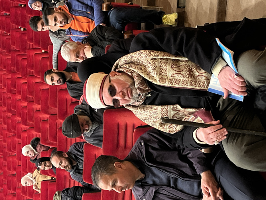 An indoor scene depicts an audience seated in an auditorium or lecture hall in the city of Rukn ad-Din Municipality, Syria. The setting features numerous rows of red upholstered seating.
In the immediate foreground, a middle-aged man with dark hair and a dark jacket is seated on the left, looking forward with his hands clasped. To his right, a man with a white beard, dark sunglasses, a black coat, and a patterned beige and brown scarf is seated. He wears a white and red striped cap. His right hand holds a dark wooden cane, and his left hand holds a blue and white booklet. He has rings on both hands. His gaze is directed slightly forward and to the left.
Behind these two individuals, various other people are seated. A woman in a black hijab is visible directly behind the prominent man. To her right, an older man with gray hair and a beard, wearing a black jacket, rests his chin on his hand. Adjacent to him is a younger man in an orange hoodie and blue puffer jacket, looking forward. Behind the man in the dark jacket, another man with a dark beard and a black beanie is visible, looking downwards.
Further into the background, multiple individuals, including men and women wearing headscarves, occupy the red seats. One man in the upper right section of the audience is seen pointing his right index finger. The lighting appears uniform, illuminating the audience from above.