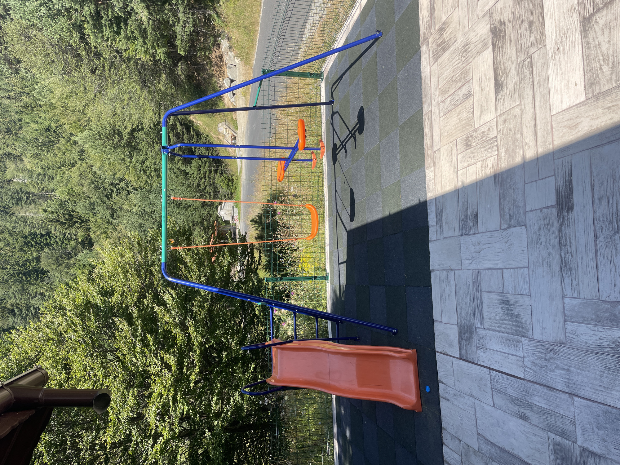 The image depicts an outdoor children's playground in a natural, mountainous setting, likely in or near Bosilkovo, Bulgaria. A blue and green metal swing set with two orange swings is centrally located. To the left of the swing set is an orange plastic slide with a blue metal frame. The playground surface consists of black and green rubber tiles, bordered by a wire fence. Beyond the fence, a paved road runs alongside the playground, with a dense forest of deciduous and coniferous trees on a hillside in the background. Sunlight casts shadows across the foreground, which is paved with light-colored, wood-patterned tiles. There are no people visible in the image.