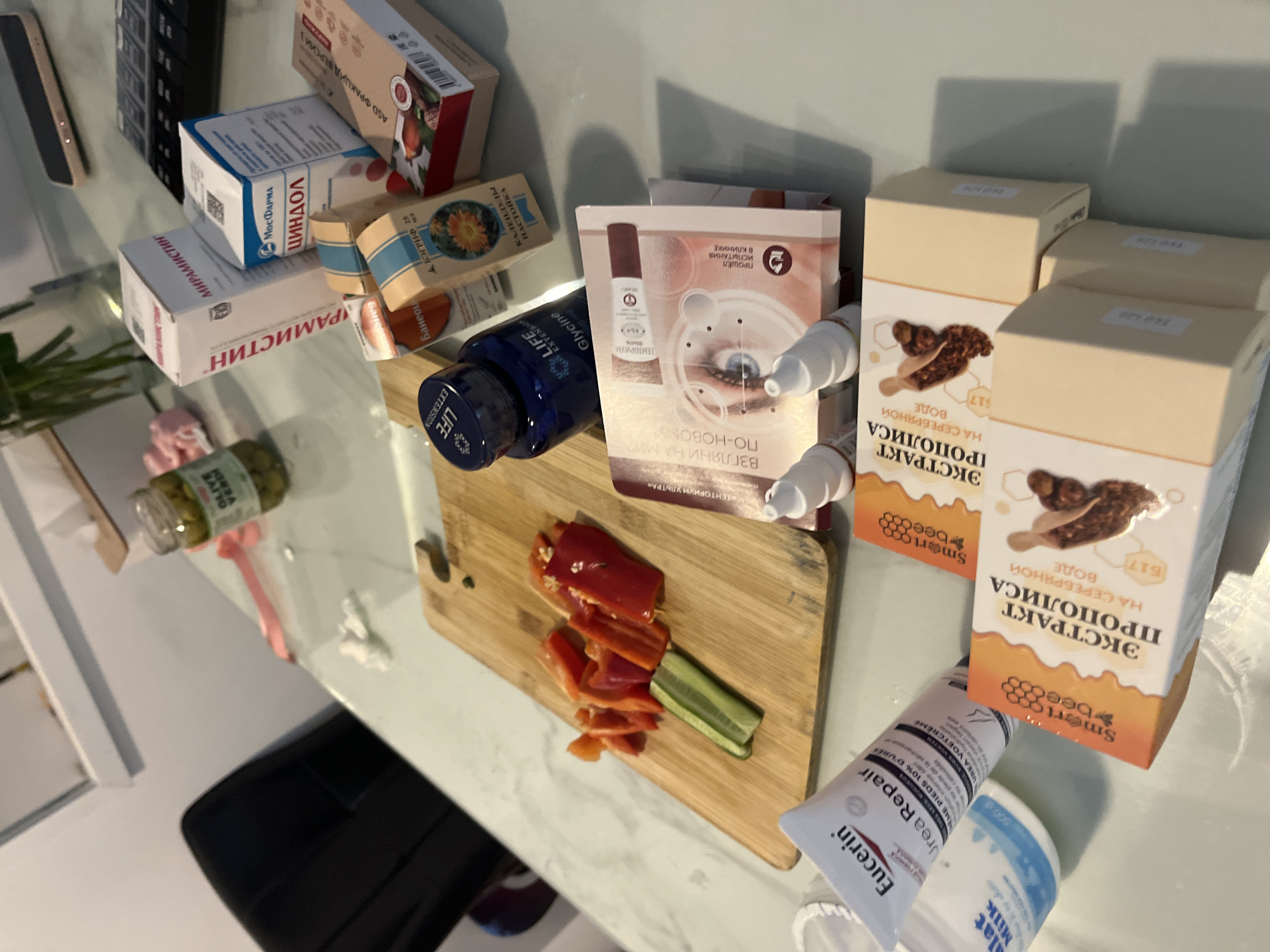 The image shows a cluttered table top with various items. There are several boxes of medications, including "МИРАМИСТИН", "ЦИНДОЛ", "ASD ФРАКЦИД ВЕРСИИ 3", and "КАЛЕНДУЛЫ НАСТОЙКА". There is a blue bottle labelled "LIFE EXTENSION", a box of "ЭКСТРАКТ ПРОПОЛИСА", a tube of "Eucerin Urea Repair", and a container of "Milk alat Milk". On a wooden cutting board, there are chopped red peppers and cucumbers. There is also a jar of olives in the background. The table is white, and a black chair is visible. There is no indication of Bac Nha Trang Ward, Vietnam.