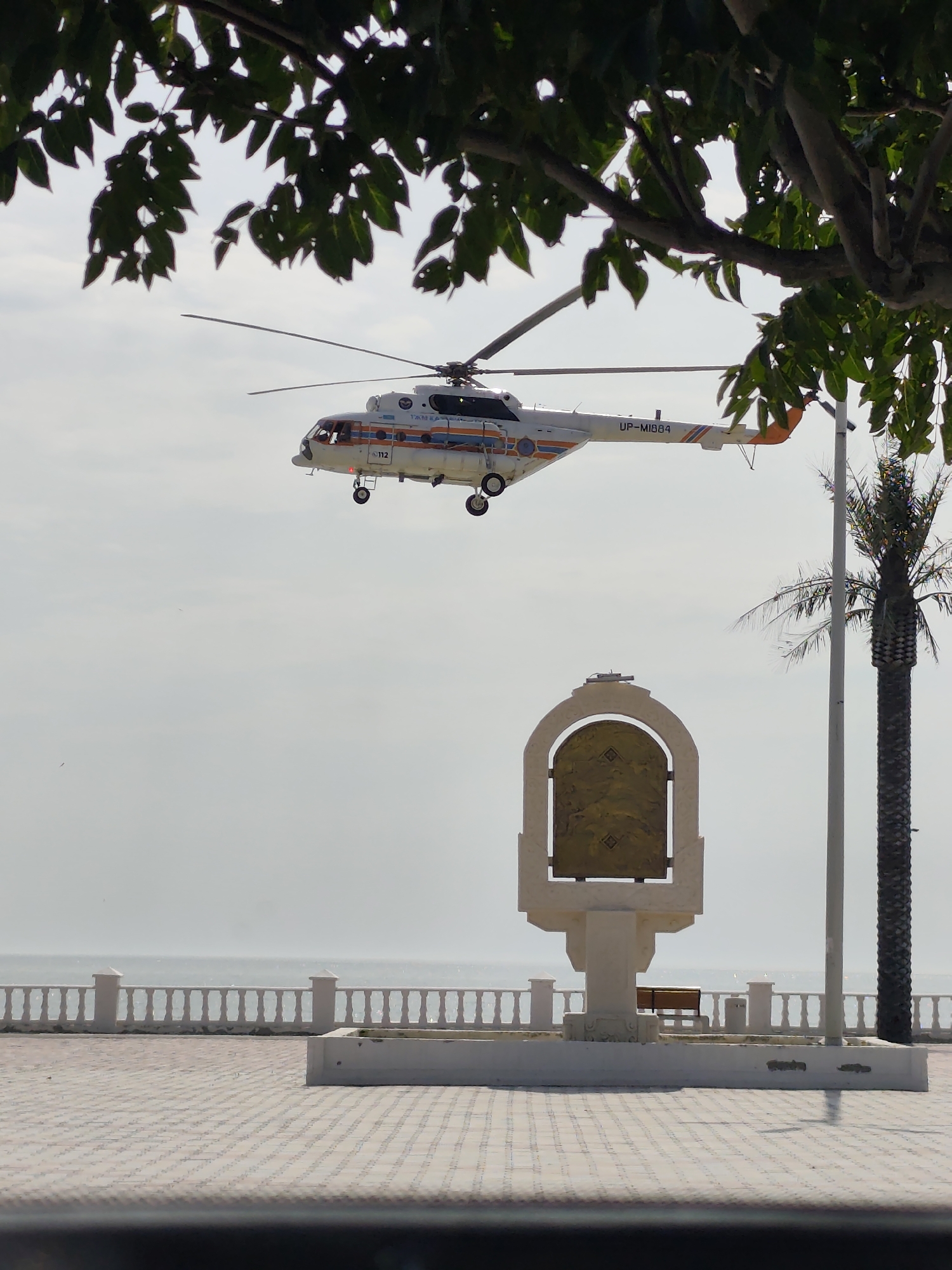 A helicopter with the registration "UP-MI884" flies over a paved area near a body of water. The helicopter is white with orange and blue accents and has "112" visible on its side. In the foreground, there is a decorative monument with an arched opening containing a gold-colored plaque. A palm tree stands to the right of the monument. Beyond the paved area is a white railing along the water's edge, and the sea is visible in the background. The scene appears to be located in Aktau, Kazakhstan, a coastal city known for its Caspian Sea promenade.