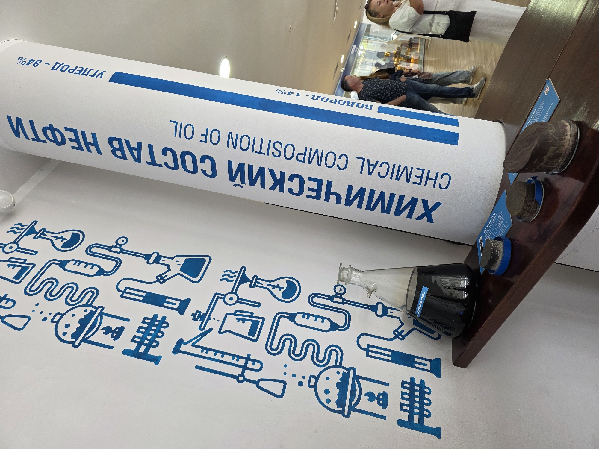 The image depicts an indoor exhibition, likely a museum or educational display, with a prominent cylindrical exhibit labeled in Russian and English. The large cylinder, oriented vertically and slightly angled towards the viewer, features text that reads "ХИМИЧЕСКИЙ СОСТАВ НЕФТИ CHEMICAL COMPOSITION OF OIL" in large blue letters. Smaller text indicates percentages, such as "УГЛЕРОД - 84%" (Carbon - 84%) and "ВОДОРОД - 14%" (Hydrogen - 14%). To the left of the cylinder, a white wall is adorned with blue, schematic drawings of laboratory equipment, including beakers, test tubes, and distillation apparatus. On a wooden shelf in the foreground, a glass flask filled with a dark liquid, presumably oil, is displayed. Two round, brown objects, possibly samples or models, rest on blue bases on the shelf. In the background, several people are visible, some looking at the exhibits. The flooring appears to be tiled. The overall impression is an educational display related to the chemical composition of oil. The presence of the text "БАЛТИЙСКАЯ НЕФТЬ" (Baltic Oil) on a label within the flask suggests a connection to the Baltic region, and given the context of Kaliningrad, Russia, this exhibit likely pertains to oil resources found in or near the Baltic Sea.