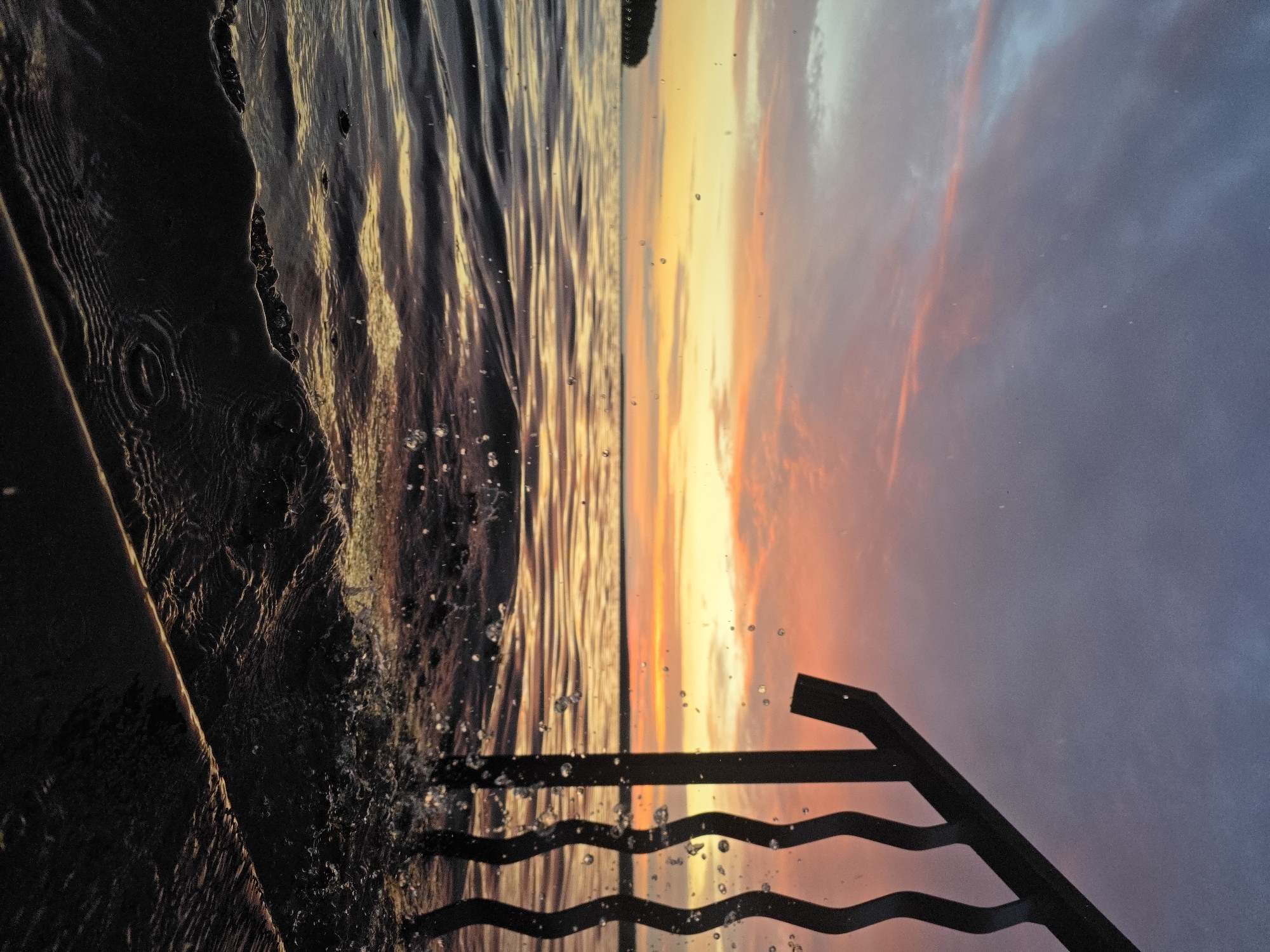A low-angle shot captures a dynamic sunset over a body of water, possibly the Kama River, near Chaikovsky, Russia. The sky is ablaze with hues of purple, pink, and orange, with wispy clouds streaking across the vibrant canvas. In the foreground, a dark, wavy railing juts out from the right side of the frame, its silhouette stark against the colorful sky. Water splashes against the railing, creating a sense of movement and energy. The water itself reflects the warm tones of the sunset, its surface rippling with gentle waves. In the distance, a dark, tree-lined shore is visible on the left horizon. The scene is devoid of people, focusing entirely on the natural beauty of the sunset and the interplay of light and water.