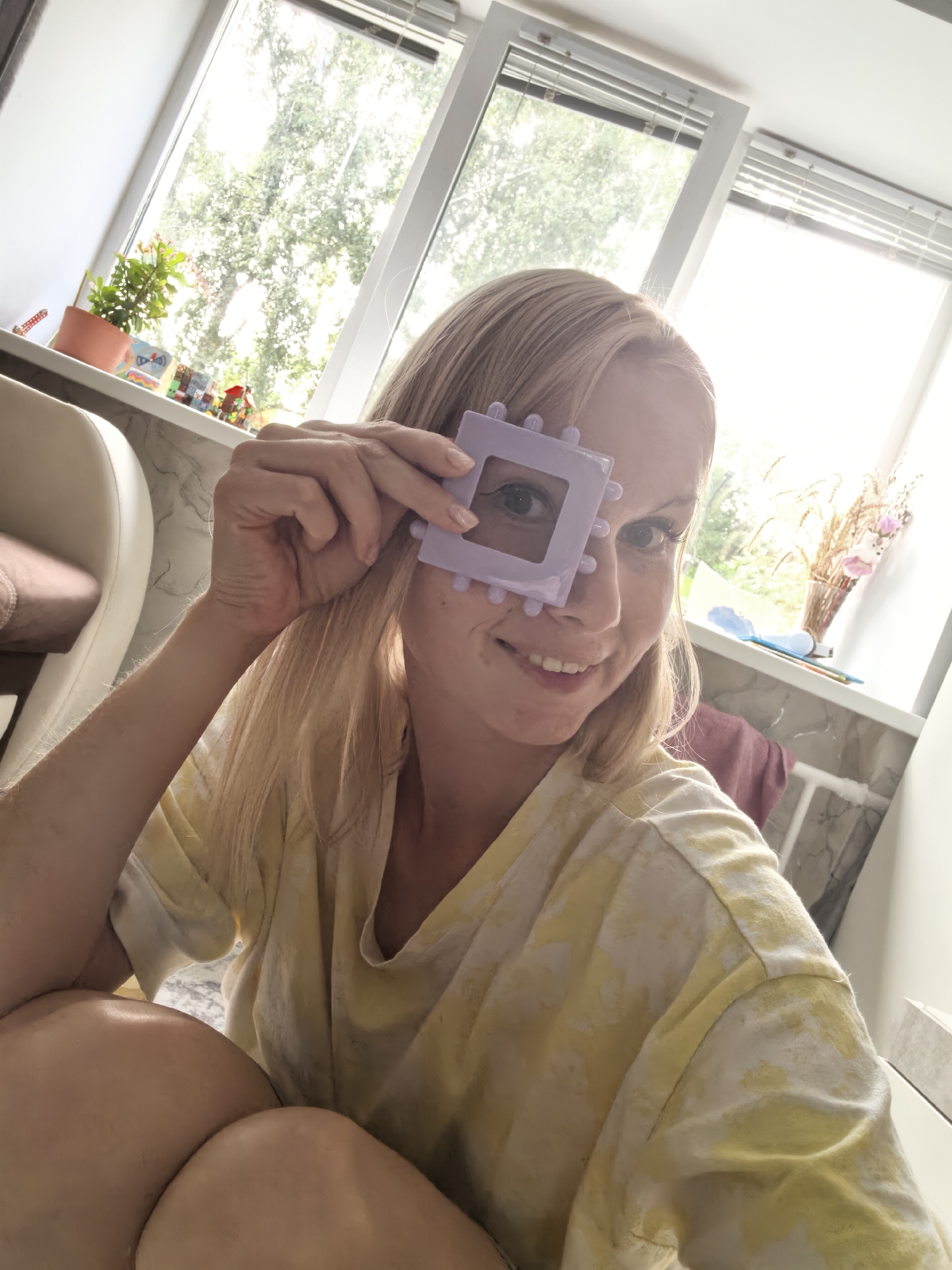 A woman with blonde hair and fair skin is sitting on the floor in front of a window. She is wearing a yellow t-shirt with a tie-dye pattern. She is holding a purple toy in front of her right eye, looking through the hole. The window is large and bright, with blinds. Outside the window, trees are visible. On the windowsill, there are several potted plants and decorative items. The room appears to be a living space. The location is identified as Sterlitamak, Russia.