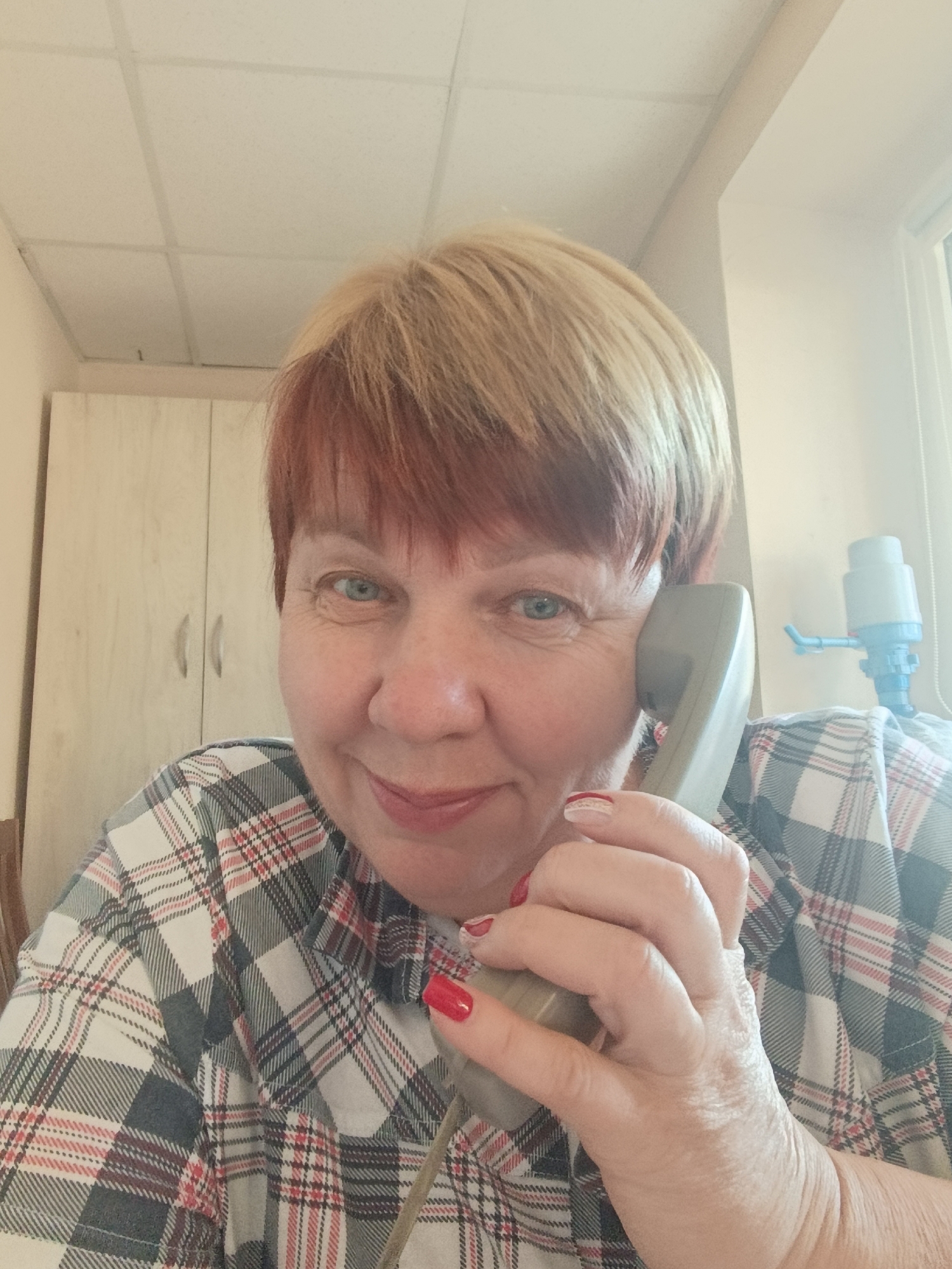 A medium shot, eye-level, indoor photograph shows a middle-aged woman with short blonde and auburn hair holding a gray telephone handset to her right ear with her right hand. The woman is looking directly at the camera with a slight smile. She has blue eyes and light skin with some freckles. She is wearing a plaid shirt with white, gray, red, and black lines. Her fingernails are painted red with a white accent on her right thumb.
The background of the image is a room with light beige walls. To the left of the woman, there are two light wood cabinets with silver handles. Above the woman's head is a white tiled ceiling. To the right of the woman, there is a window, and a blue and white water dispenser is visible on a shelf. The overall lighting suggests an office or administrative setting, potentially in Svietlahorsk, Belarus, given the context.