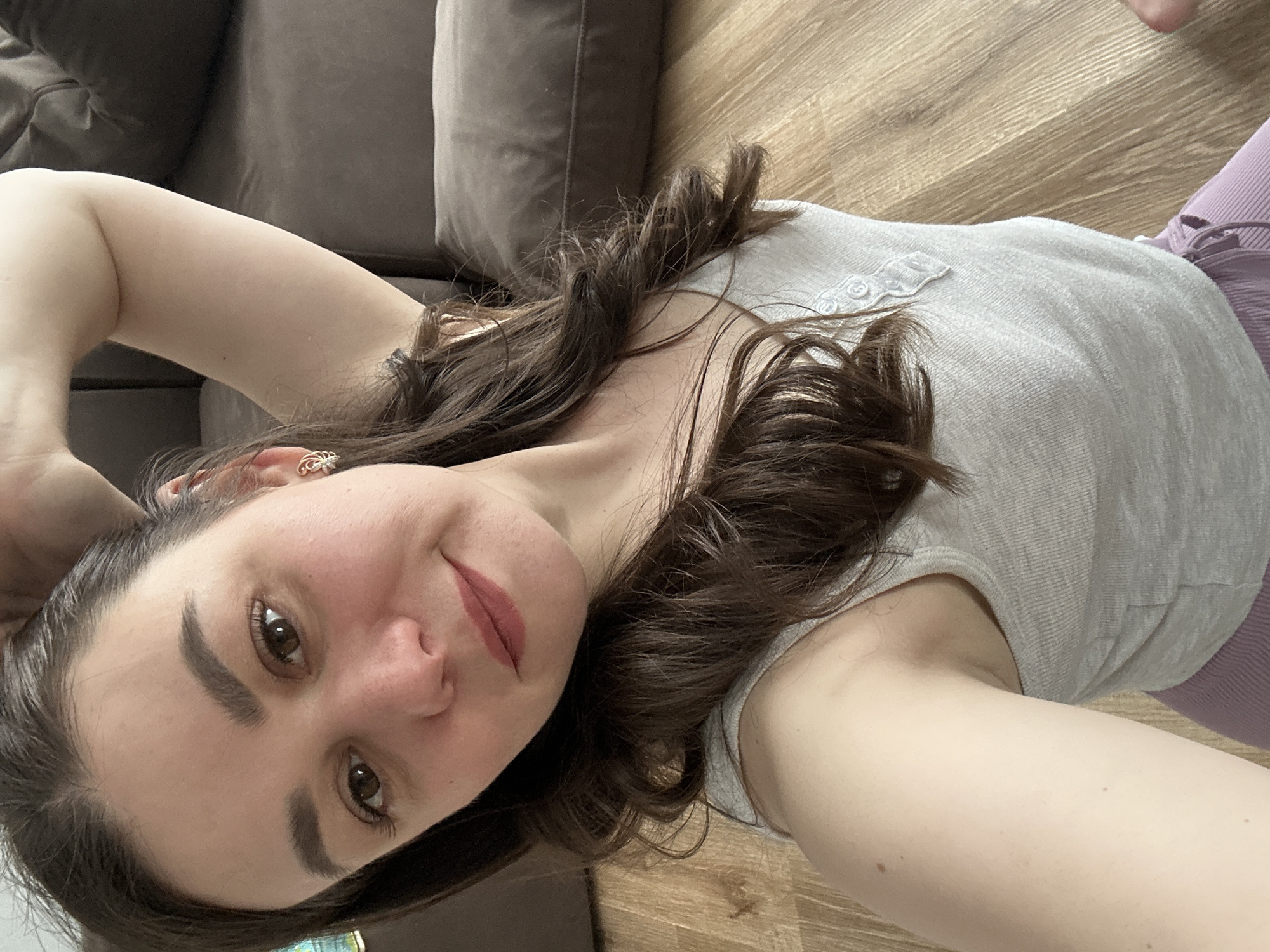 A woman is lying on a wooden floor next to a brown couch. She is wearing a gray shirt with small buttons and purple leggings. Her brown hair is styled with loose waves. She has brown eyes, fair skin, and is wearing lipstick. Her right hand is behind her head. The exact location within the city of Chaikovsky, Russia, cannot be determined from this image.
