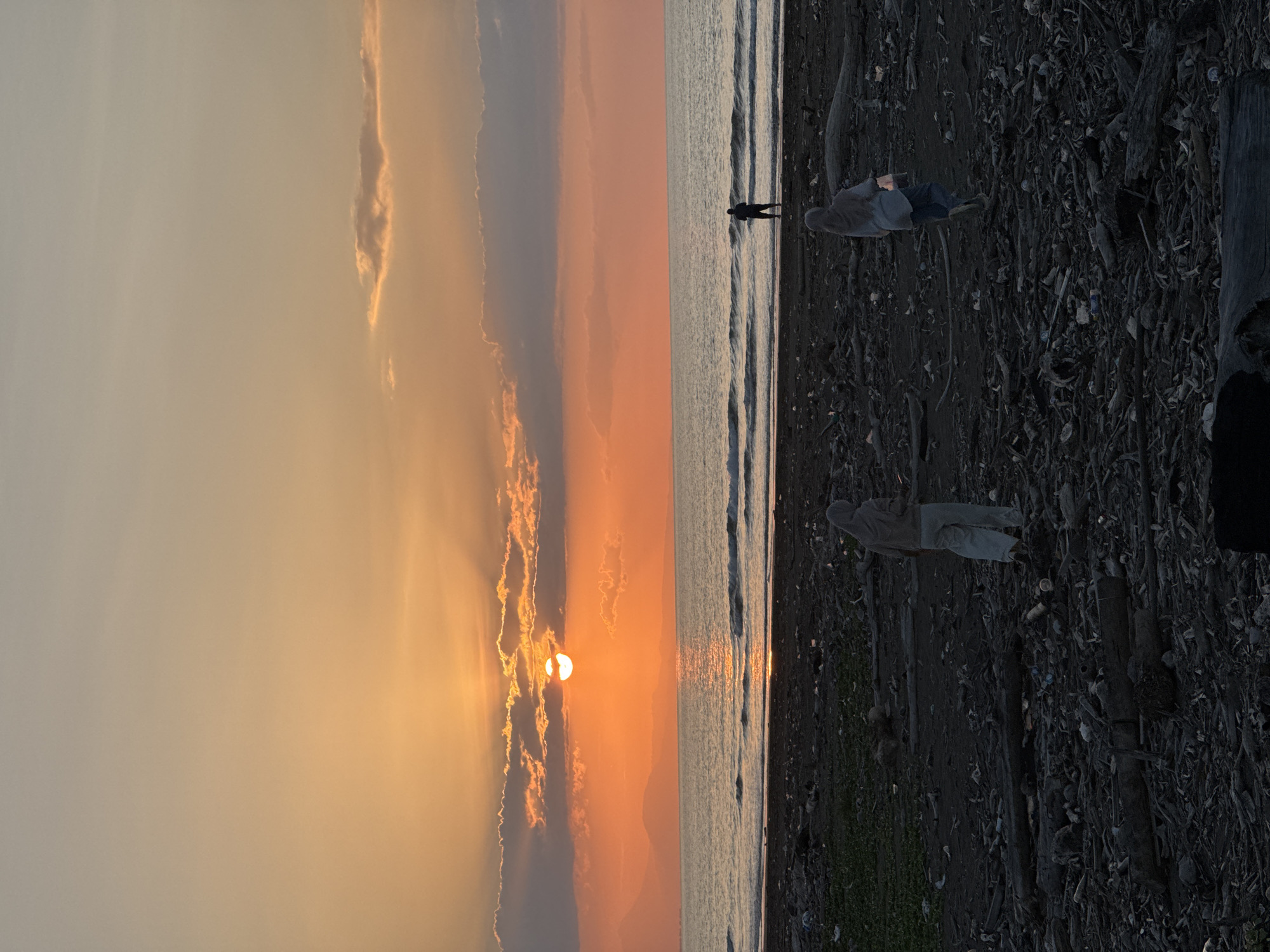 A wide shot captures a sunset over a beach in Meunasah Jurong, Indonesia. The sun, a bright orange orb, is partially obscured by clouds, casting a warm glow across the sky and reflecting on the calm sea. The foreground is a dark, debris-strewn beach, with scattered driftwood and litter. Two figures, dressed in casual clothing and headscarves, walk across the beach in the foreground, facing away from the camera. One figure is closer to the viewer and is holding a small lantern. In the middle ground, a lone figure stands at the water's edge, facing the ocean. The overall scene depicts a tranquil yet slightly somber end to the day.