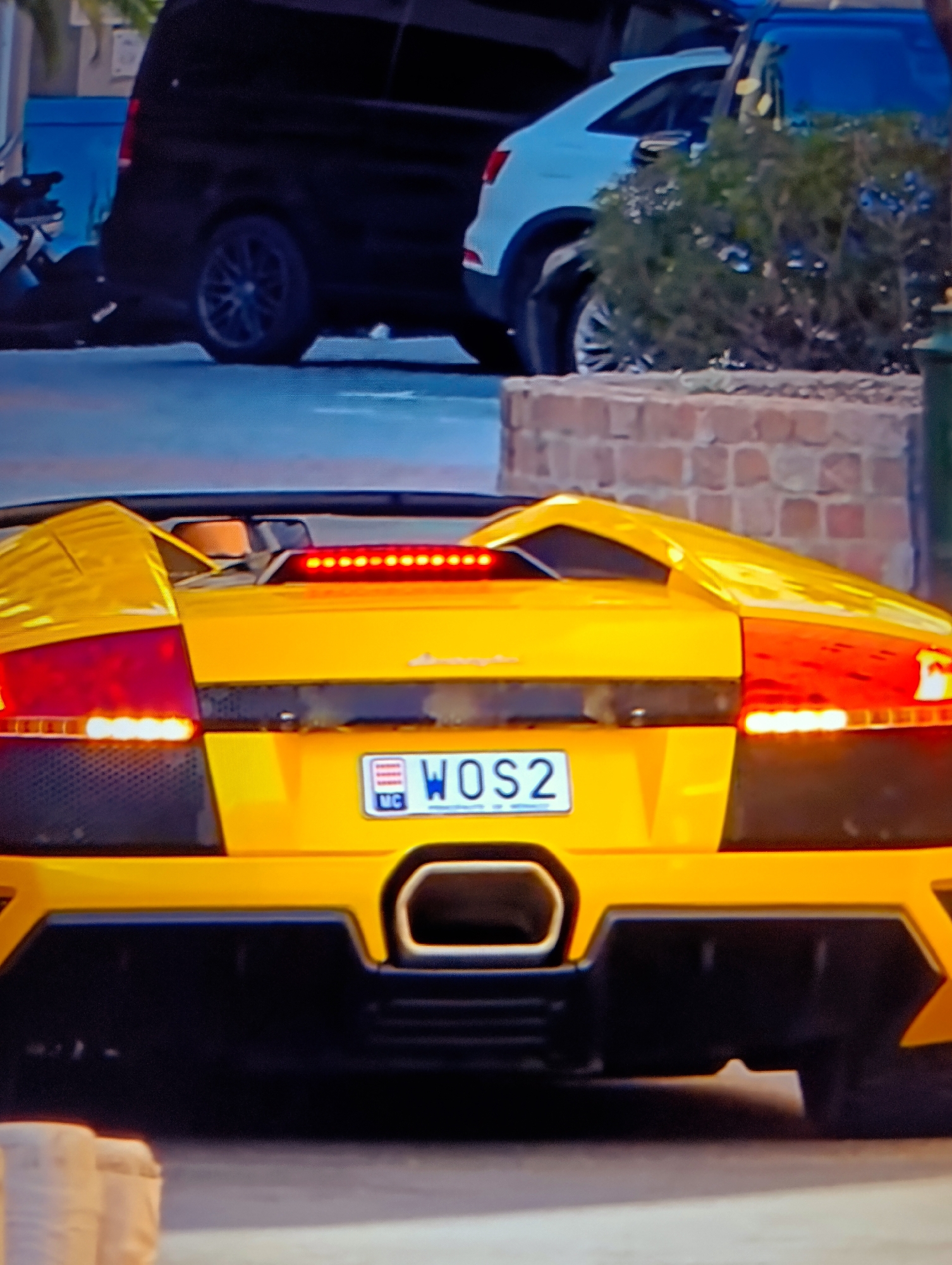 A yellow Lamborghini is parked on a street in an urban setting, with other vehicles and foliage visible in the background. The license plate reads "WOS2" and is marked with "MC," suggesting it is from Monaco. The exact location within the city is indeterminable based solely on the visual information provided.