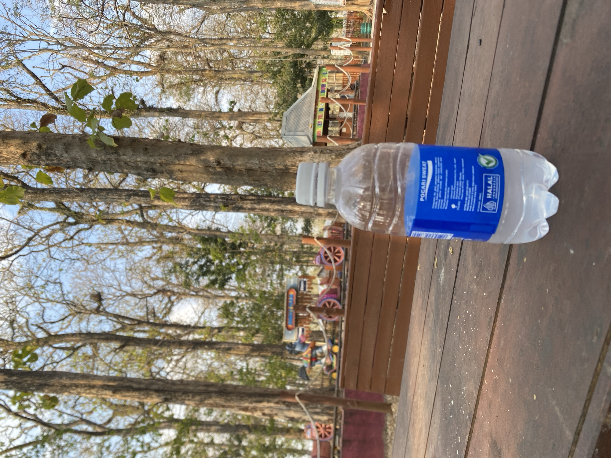 A partially filled plastic bottle of Pocari Sweat sits on a wooden table in an outdoor setting. The bottle's blue and white label is visible, featuring text and a halal certification. In the background, a wooden fence separates the seating area from a play area. Behind the fence, a small train-like amusement ride with colorful cars is set up. Tall trees with sparse foliage form a canopy overhead, and the sky is visible through the branches. The environment appears to be a park or recreational area, possibly located in Ketileng, Indonesia, given the halal certification on the bottle. The lighting suggests it is daytime, with sunlight casting shadows on the table. There are no people visible in the immediate foreground.