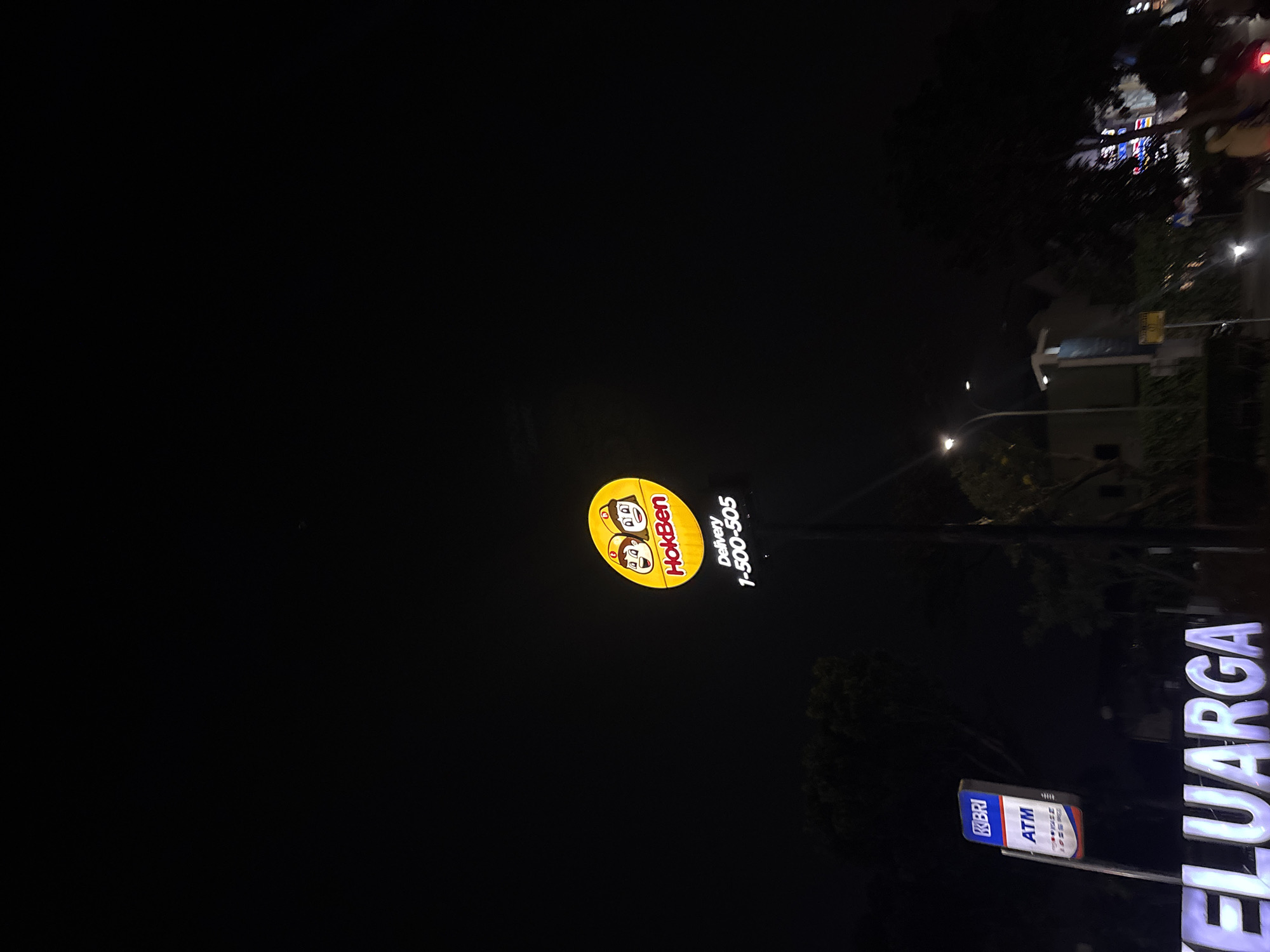 This media file depicts a nighttime scene in South Tangerang, Indonesia. The primary subjects are illuminated signs. A prominent, circular yellow sign features the "HokBen" logo, which includes two cartoon characters and the text "HokBen" and "Delivery 1-500-505". To the right of this, a large, white, illuminated sign displays the word "KELUARGA" in capital letters. Below the HokBen sign, a smaller blue and white sign indicates "BRI ATM". The setting appears to be a street or commercial area at night, with streetlights visible in the background and the ambient darkness suggesting it is after sunset. There are no people clearly visible. The overall mood is that of an urban commercial landscape at night, with the bright lights of the signs contrasting with the dark surroundings.