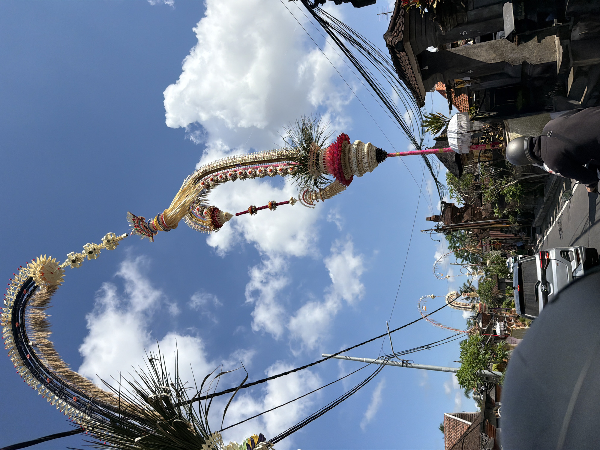 The image depicts a street scene in Tampaksiring, Indonesia, likely during a cultural or religious festival. The most prominent features are two elaborately decorated, towering arches adorned with flowers, leaves, and straw-like materials. These arches are characteristic of Balinese decorations, possibly for Galungan or other local celebrations.
In the foreground, a person wearing a black jacket and a grey helmet rides a motorcycle. The motorcycle is in motion, indicating that the photo was taken from a moving vehicle. Behind the motorcycle, a silver SUV is visible, also appearing to be in traffic.
The street is lined with traditional Balinese architecture, featuring tiled roofs and stone carvings. Several utility poles with a complex network of wires are also present, suggesting a developed urban or semi-urban area. The sky is a bright blue with scattered white clouds, indicating fair weather. The overall impression is of a vibrant, culturally rich environment amidst everyday life.