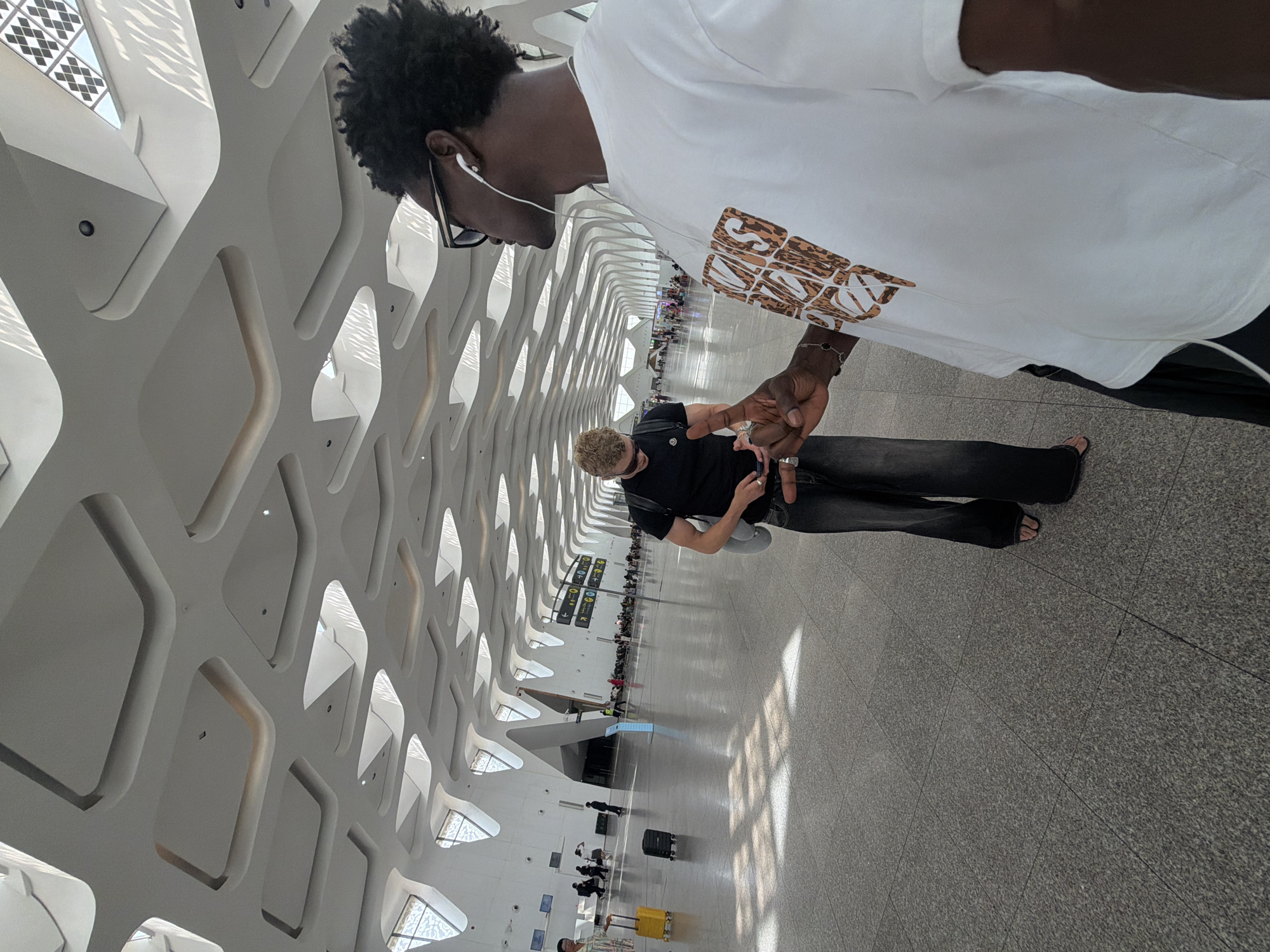 The image shows two young men in a modern airport terminal. The man in the foreground, facing right, is wearing a white t-shirt with a graphic print, sunglasses, and white earbuds. He is gesturing with his right hand, index finger extended. The man in the background, facing left and partially obscured, is wearing a black t-shirt and dark, wide-leg jeans. He is looking at his phone. The terminal features a distinctive, undulating white ceiling with integrated lighting and skylights, characteristic of Marrakech Menara Airport's architectural design. The floor is polished stone, and the overall environment appears clean and spacious, with signs and other travelers visible in the distance. Sunlight streams through the skylights, creating geometric patterns of light and shadow on the floor.