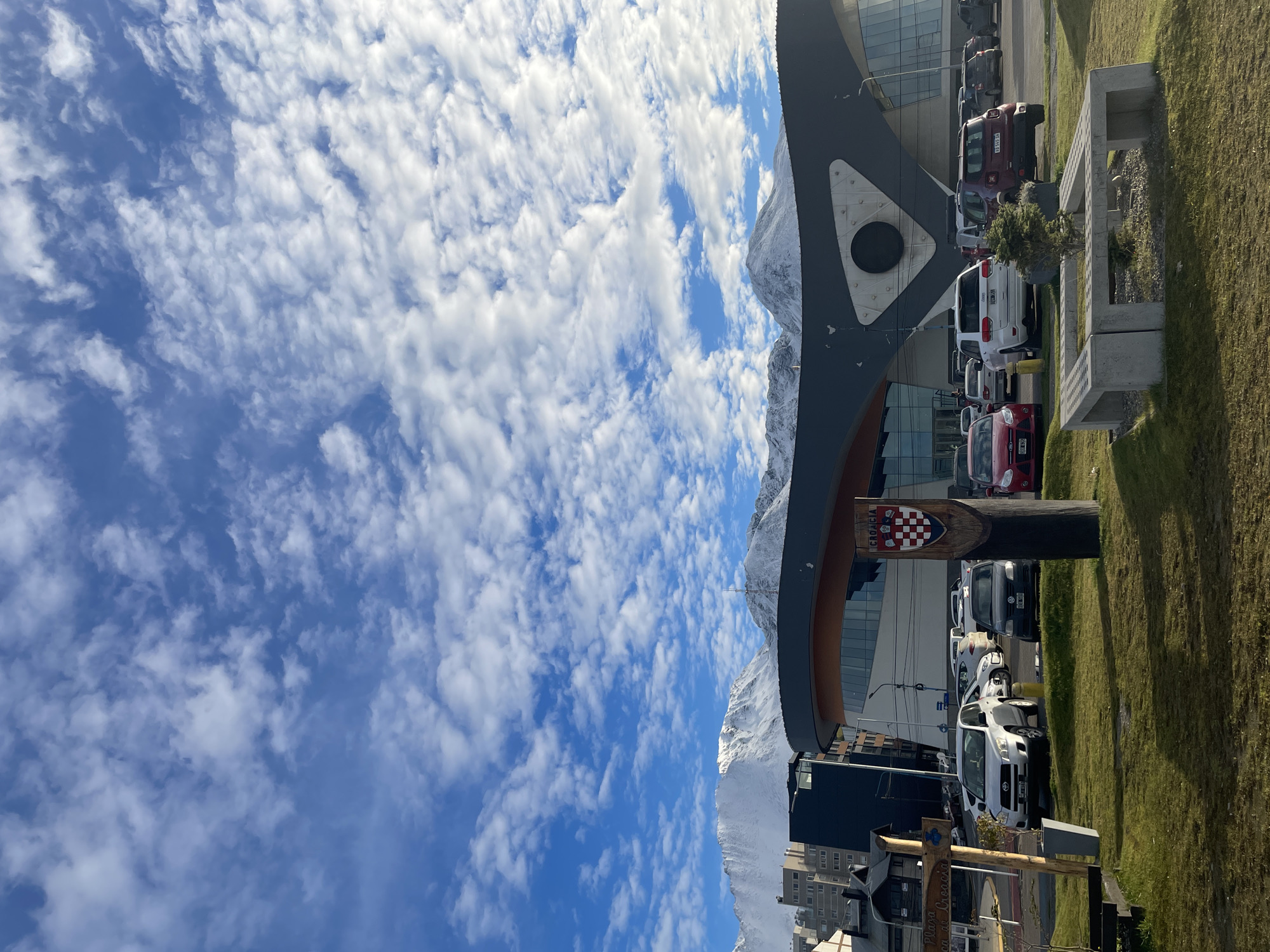 The image captures a scene in Ushuaia, Argentina, showcasing a dynamic interplay of urban architecture and natural landscape. The prominent element is a modern building with a curved design and a dark gray facade featuring a circular detail. The building is partially obscured by a tall, wooden structure bearing the Croatian national flag and the word "CROACIA," indicating a Croatian cultural or community presence. Behind the building and structure are snow-capped mountains, which dominate the skyline. The sky is bright blue with numerous white, fluffy clouds. Adjacent to the building, there is a parking area filled with a variety of vehicles, mainly cars and vans. A patch of green grass and a concrete bench are located at the edge of the parking area. In the foreground, there are building roofs and street signs, partially obscured.
